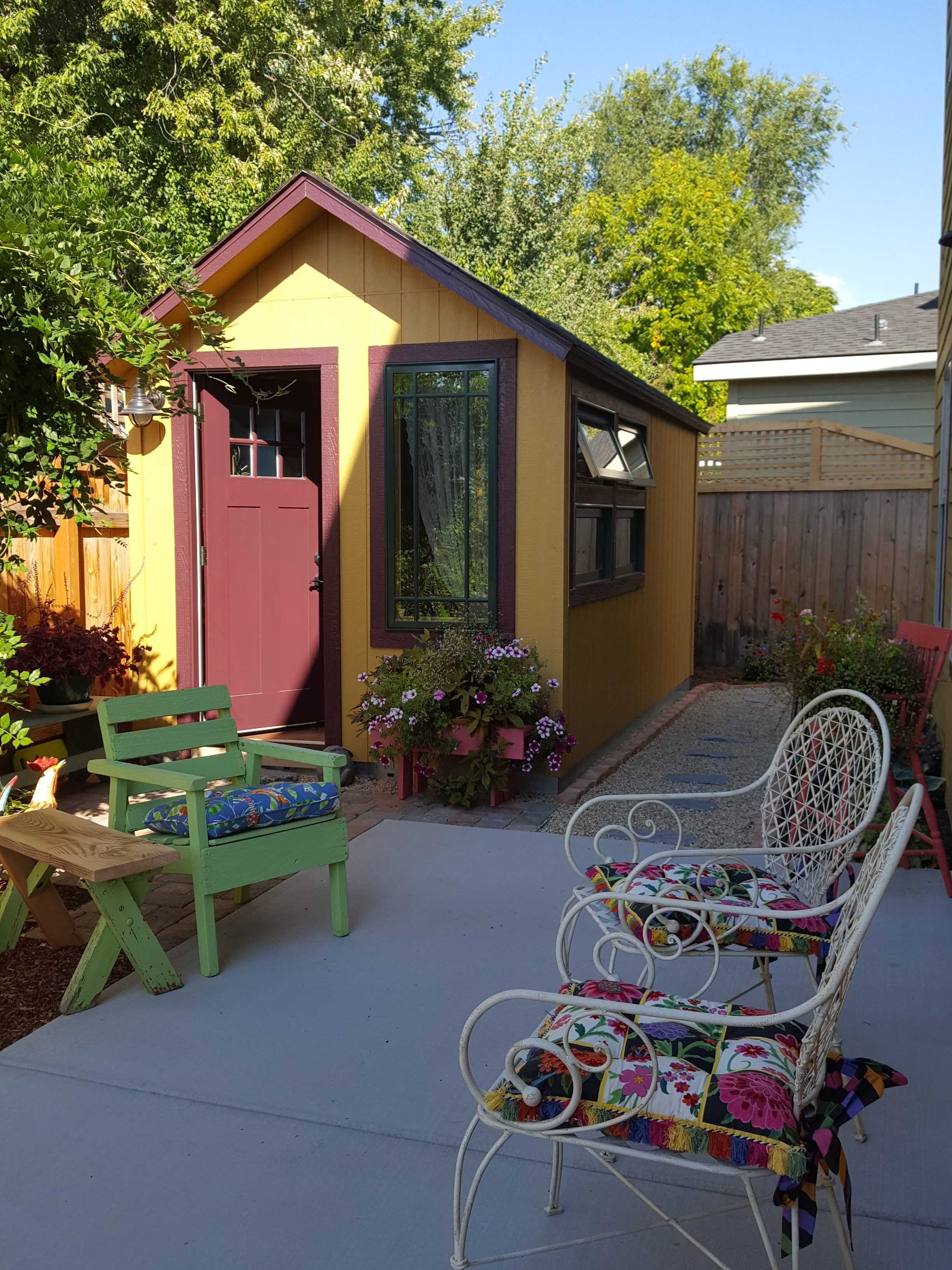 A small, colorful garden shed with a red door and window, surrounded by vibrant flowers and patio furniture on a sunny day.
