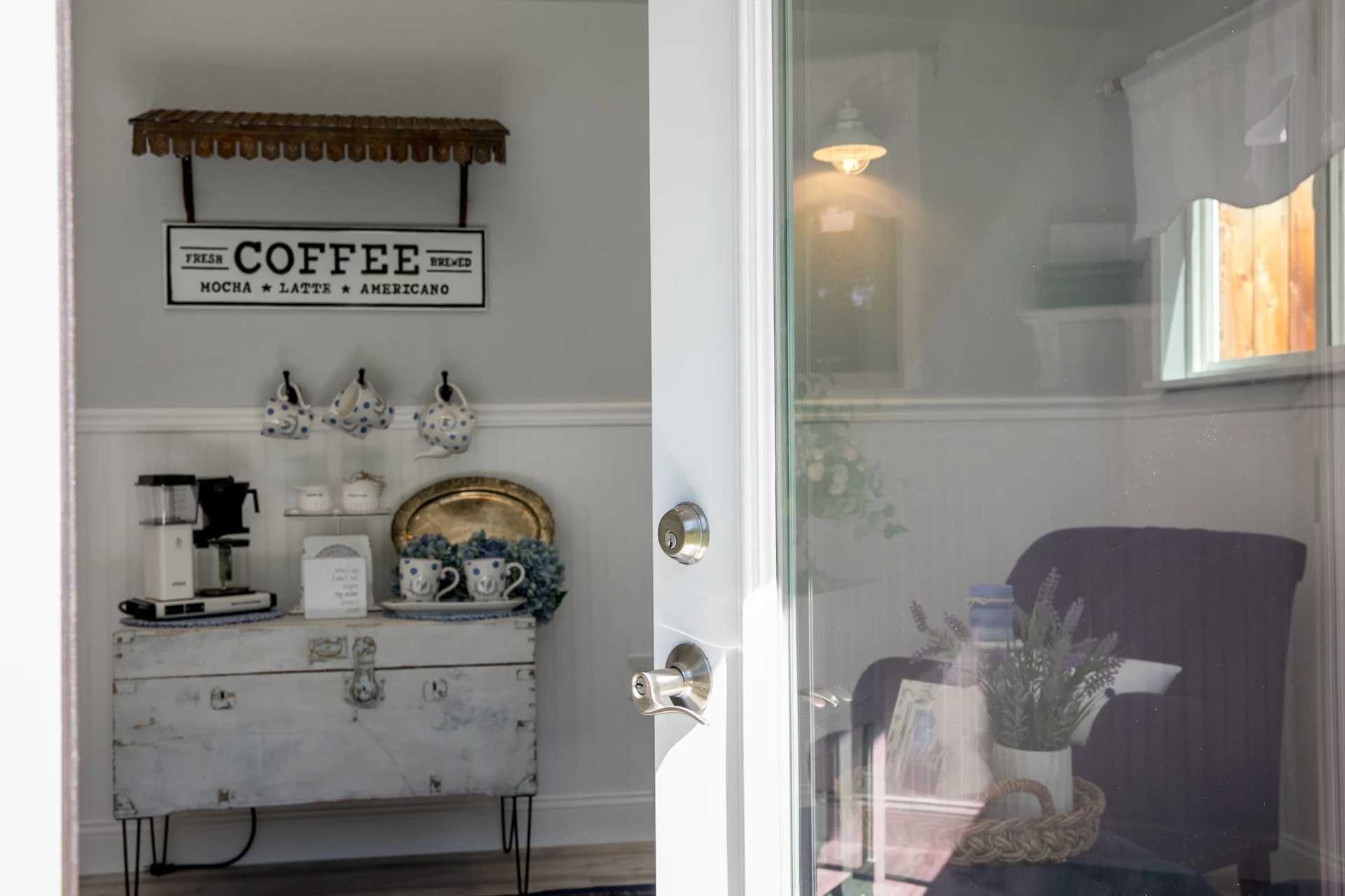 A cozy coffee nook with mugs, a coffee machine, and decor on a white cabinet. A reflection shows a chair and lamp.