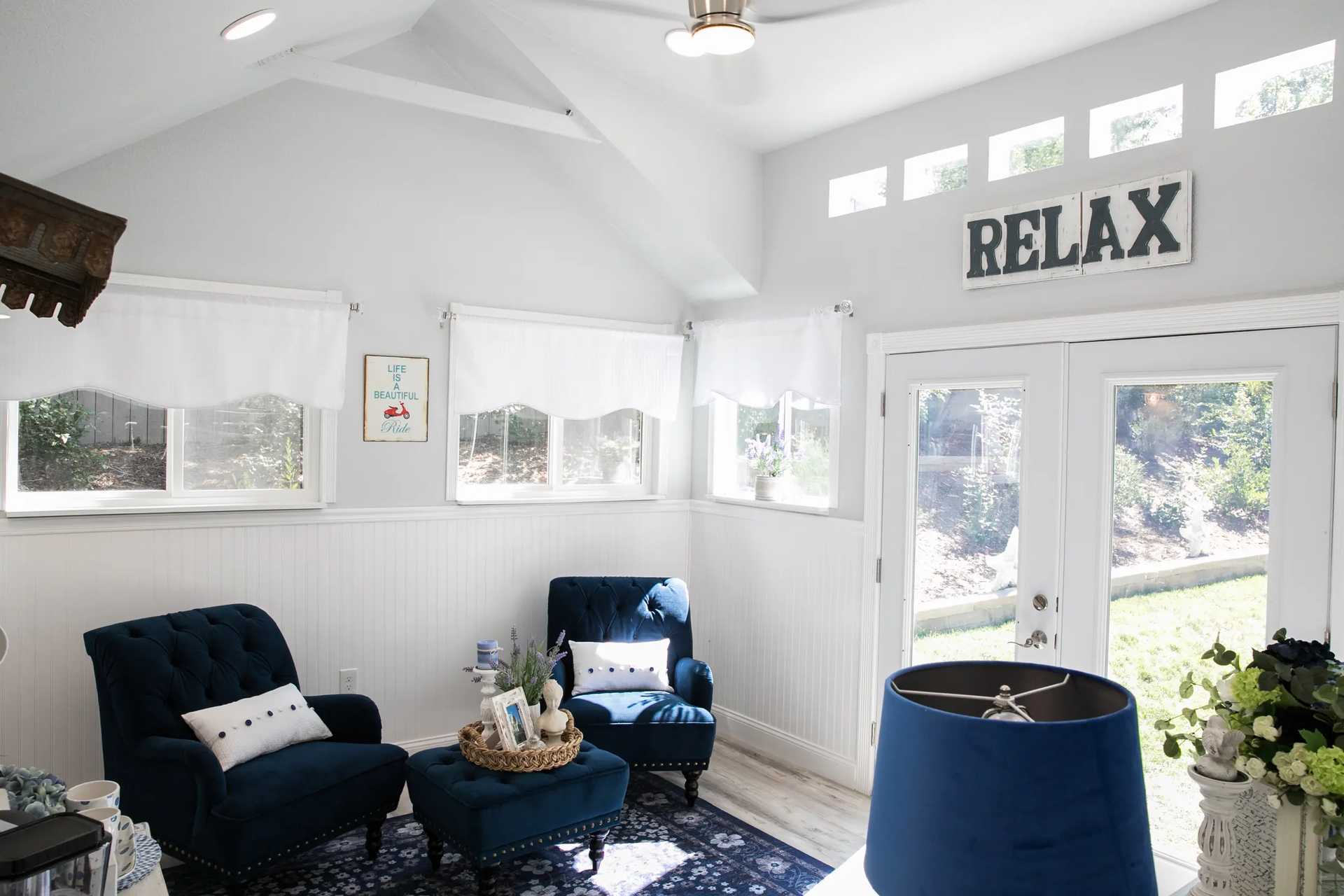 Bright sunroom with blue chairs and accents, white curtains, and a "Relax" sign above glass doors leading to a garden view.