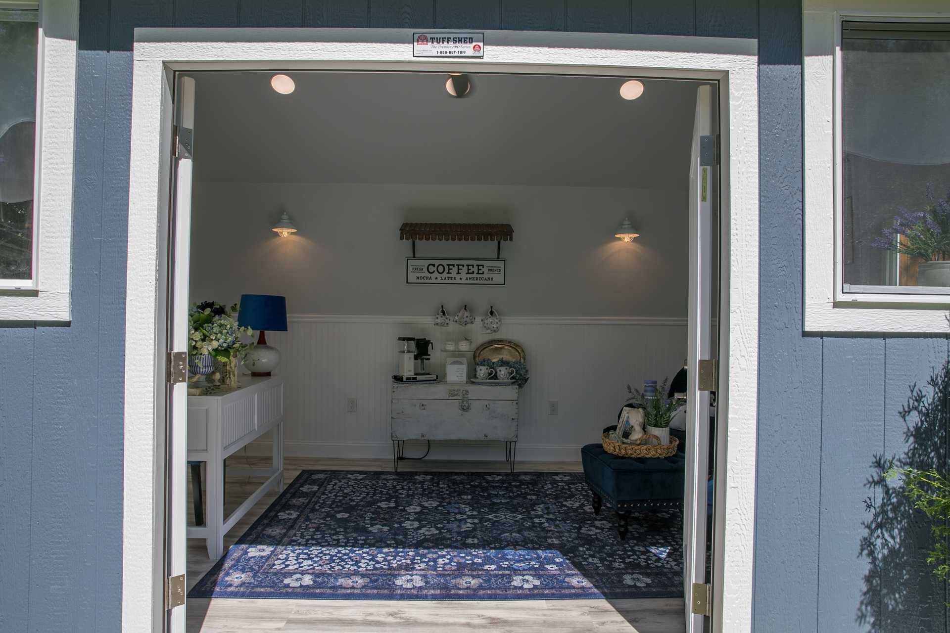 Linda's Interior TR-800 She Shed Open door view of a cozy room with a coffee station, blue rug, white furniture, and decorative plants. Sign reads "COFFEE" on the back wall.