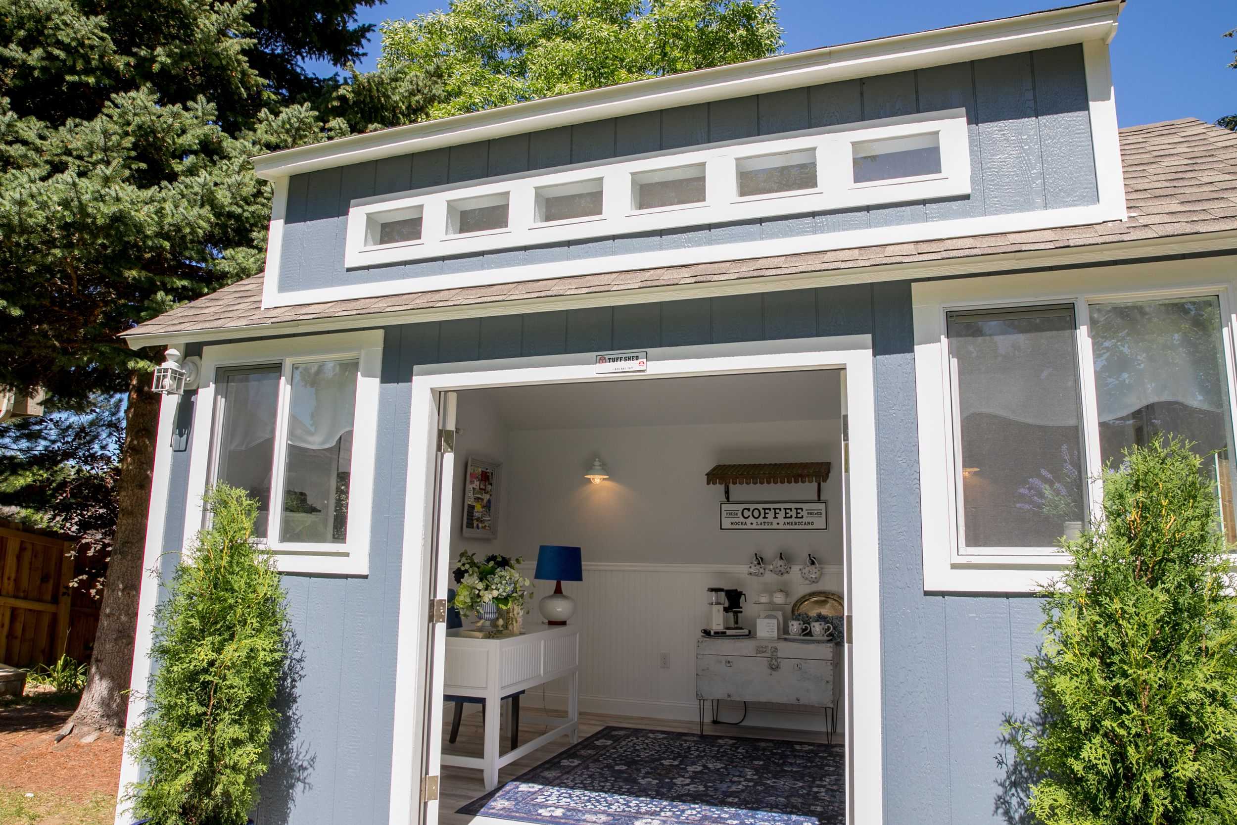 TR800 shed with white trim, open doors revealing a cozy interior with a coffee station, lamp, and rug. Surrounded by trees and greenery.