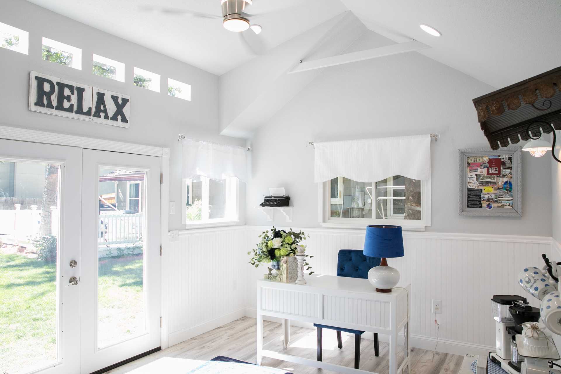 Linda's Interior TR-800 She Shed 1 Bright room with white walls, a desk with a blue lamp, flowers, and a "RELAX" sign. Large windows and doors let in natural light.