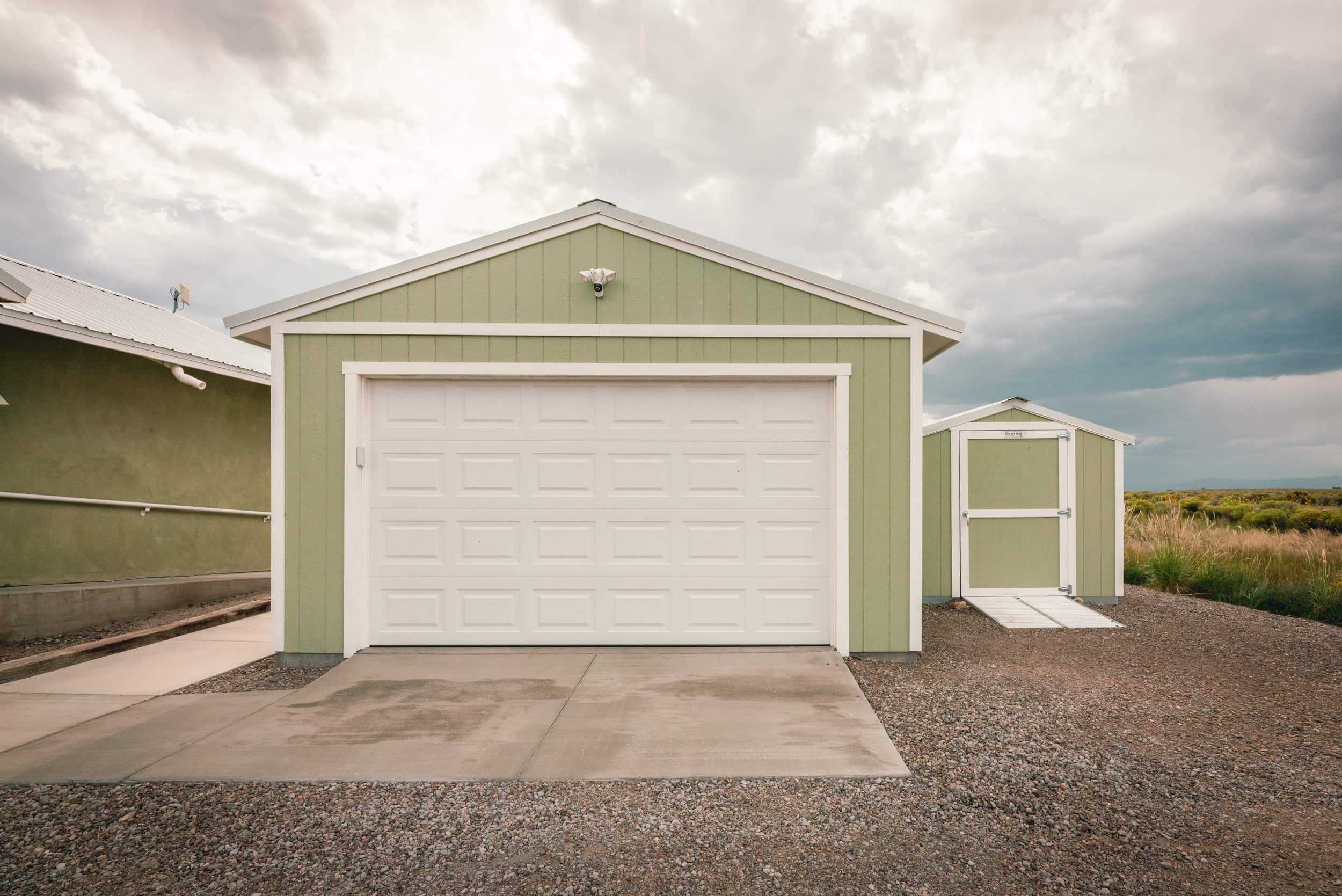 A green Sundance Ranch Garage with a white door and a small matching shed on a gravel driveway