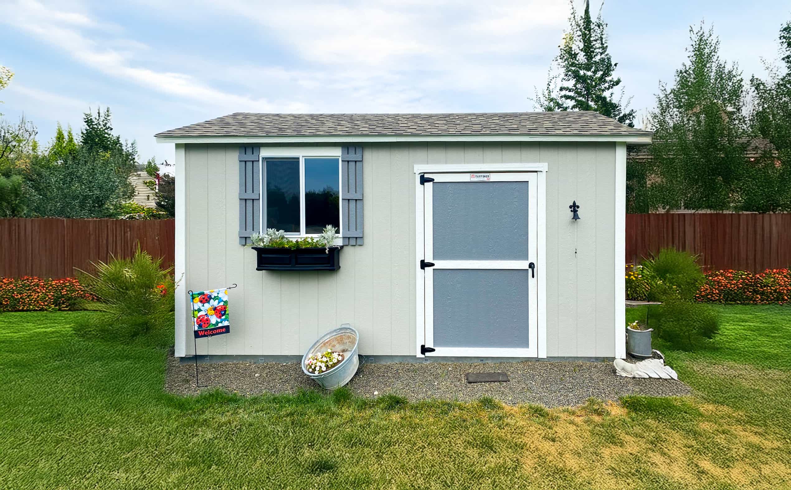 A TR800 gray shed with a window box of plants, a welcome flag, and a metal tub with flowers, set in a grassy backyard.
