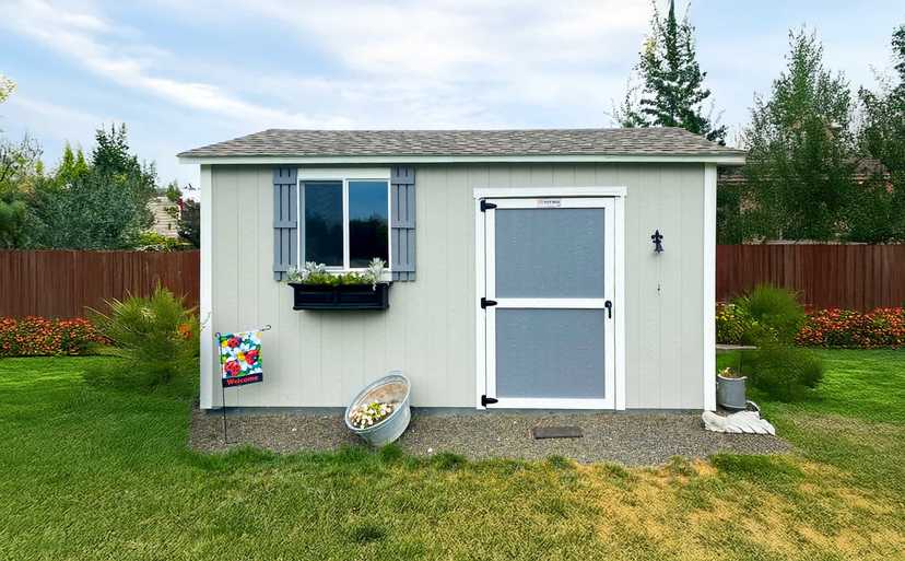 A TR800 gray shed with a window box of plants, a welcome flag, and a metal tub with flowers, set in a grassy backyard.