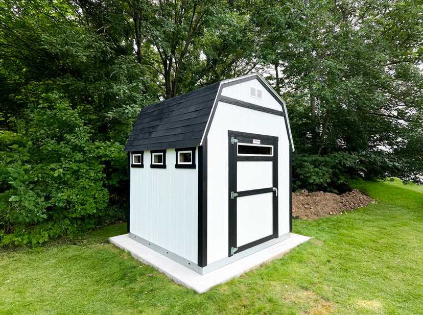 White and black barn on a cement pad against a backdrop of dark green trees
