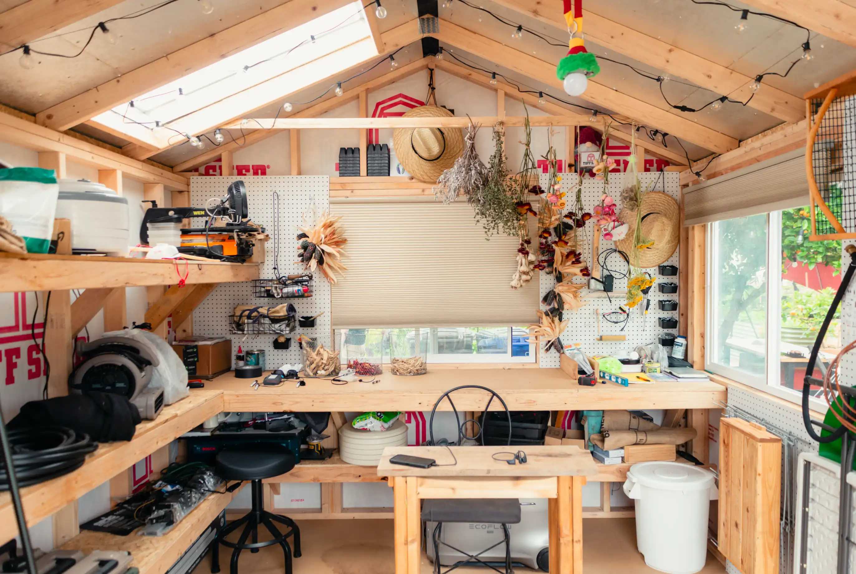 A tidy workshop with wooden shelves, tools, hats, and decorations. Central table and chairs under a skylight, surrounded by organized supplies.