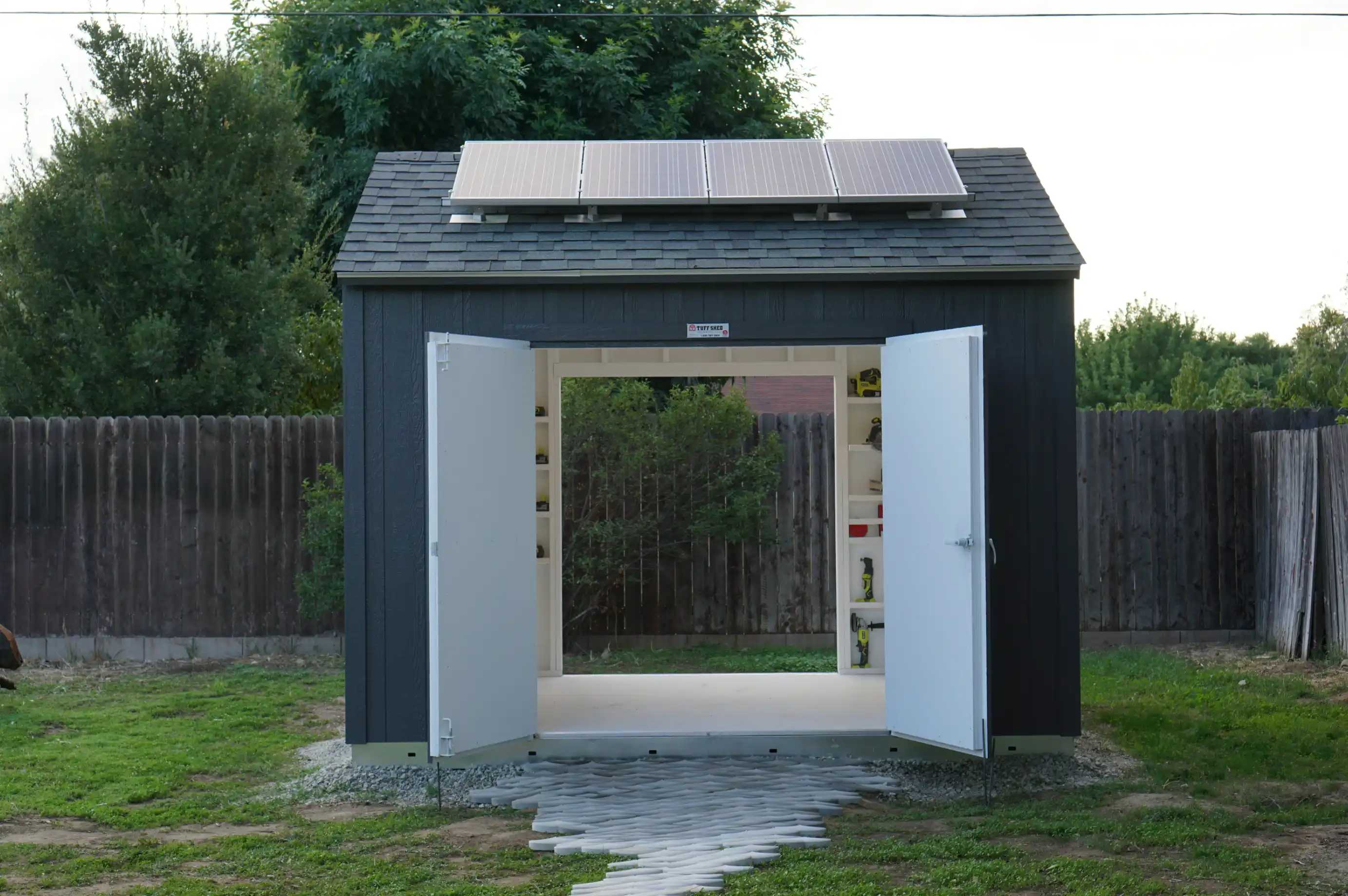 A small dark-colored shed with open double doors and solar panels on the roof, set in a grassy yard with a wooden fence and trees in the background.