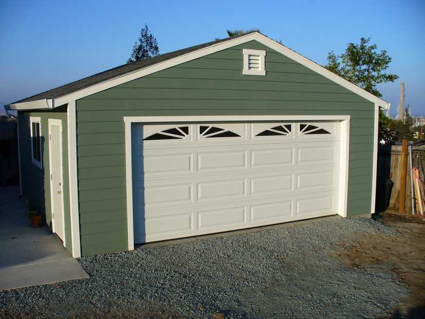 A green garage with a white door and a gravel driveway, set in a backyard with trees and a clear blue sky in the background.
