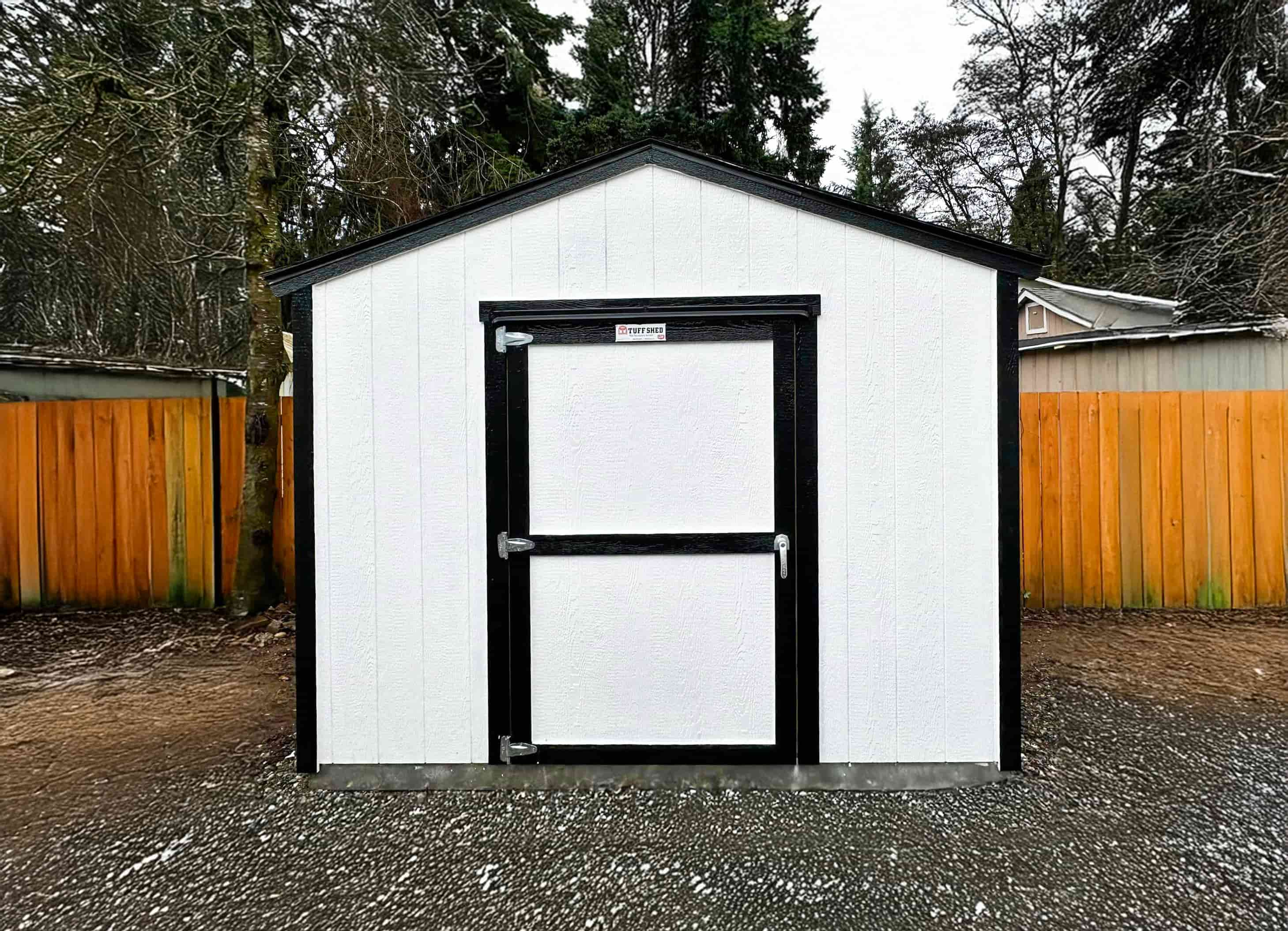 A white TR700 shed with a black-trimmed door and roof stands on a gravel surface.