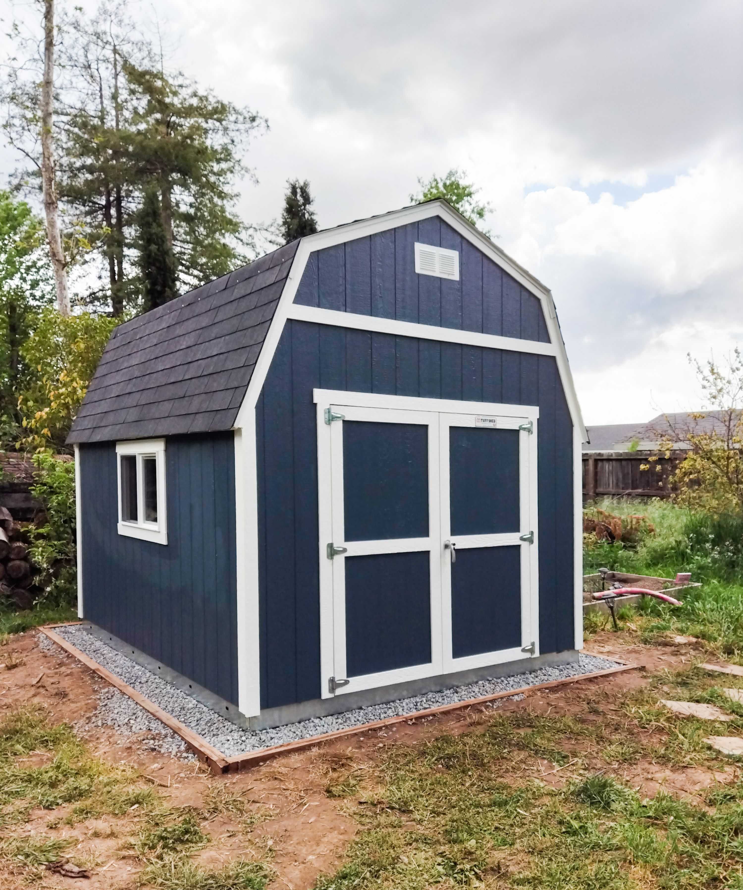 A blue TB600shed with white trim and a gabled roof stands on a gravel base in a grassy backyard