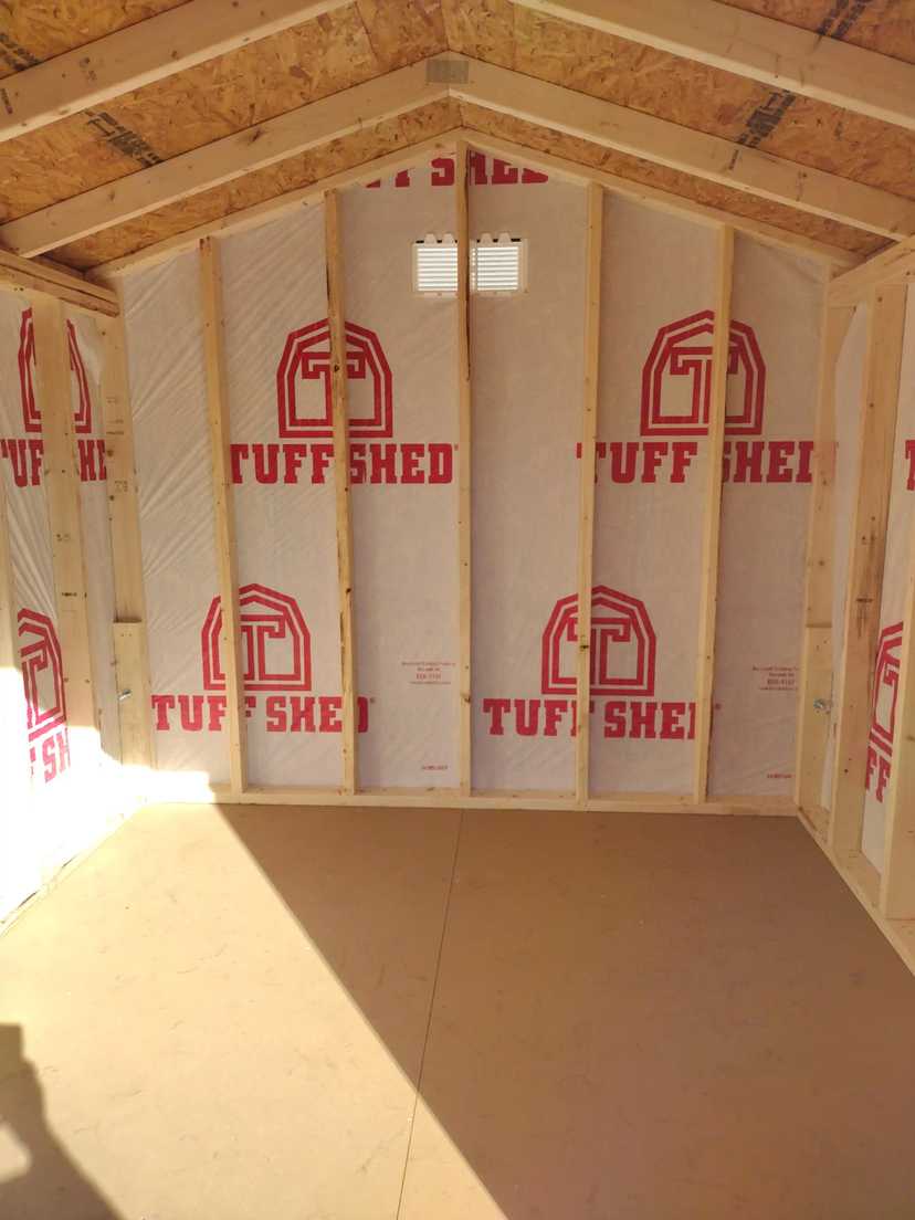 Interior view of a wooden shed under construction, with visible insulation marked "Tuff Shed" and sunlight streaming in from the left.