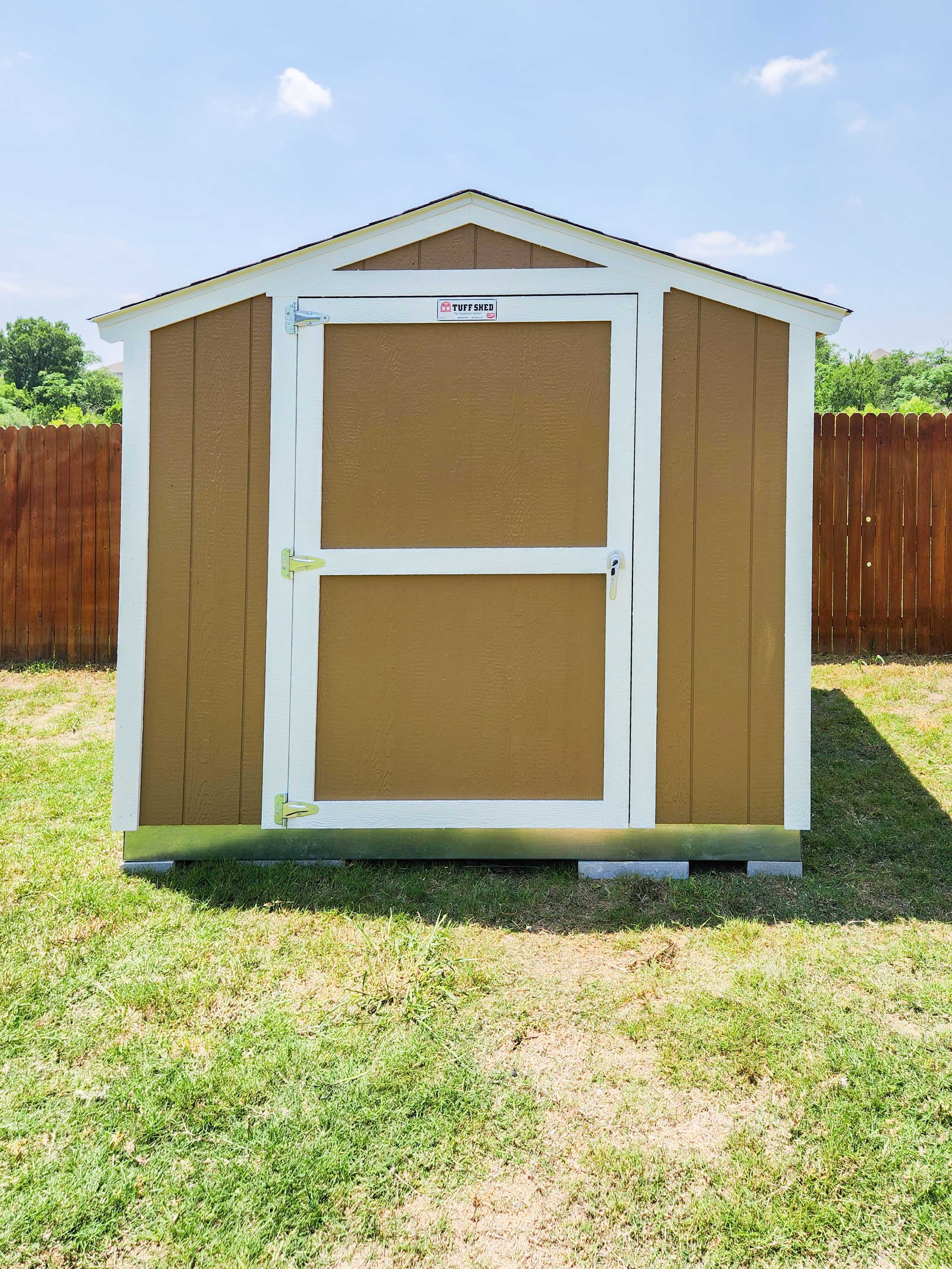 A SR600 brown and white shed with a closed door, set in a grassy yard with a wooden fence and trees in the background under a clear sky.
