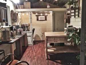 Home Office Shed Cozy, rustic home office with wooden furniture, plants, a large desk, shelves, and a small seating area. Warm lighting creates a welcoming ambiance.