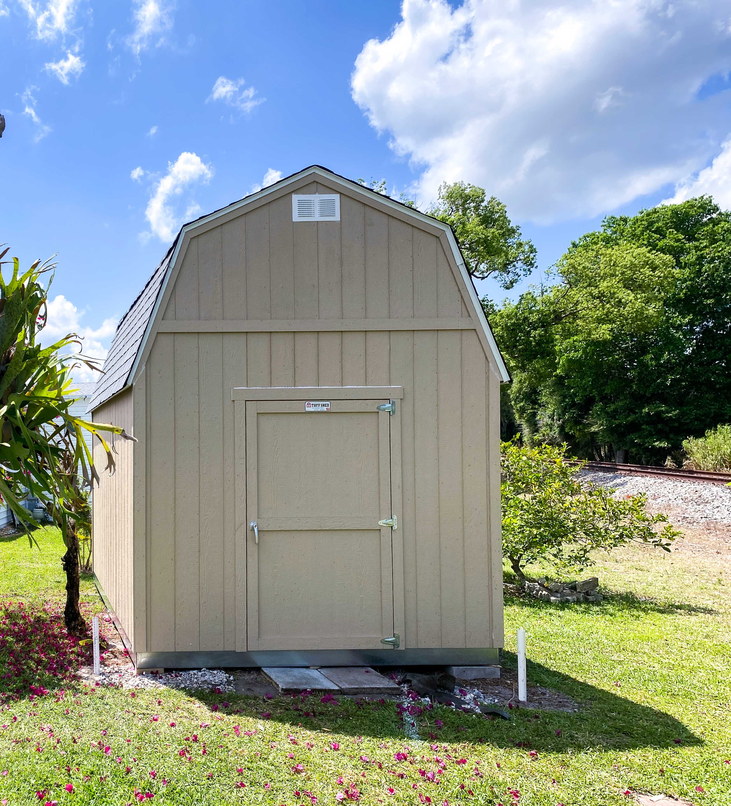 A TB700 beige shed with a gabled roof stands in a grassy area near a railroad track.