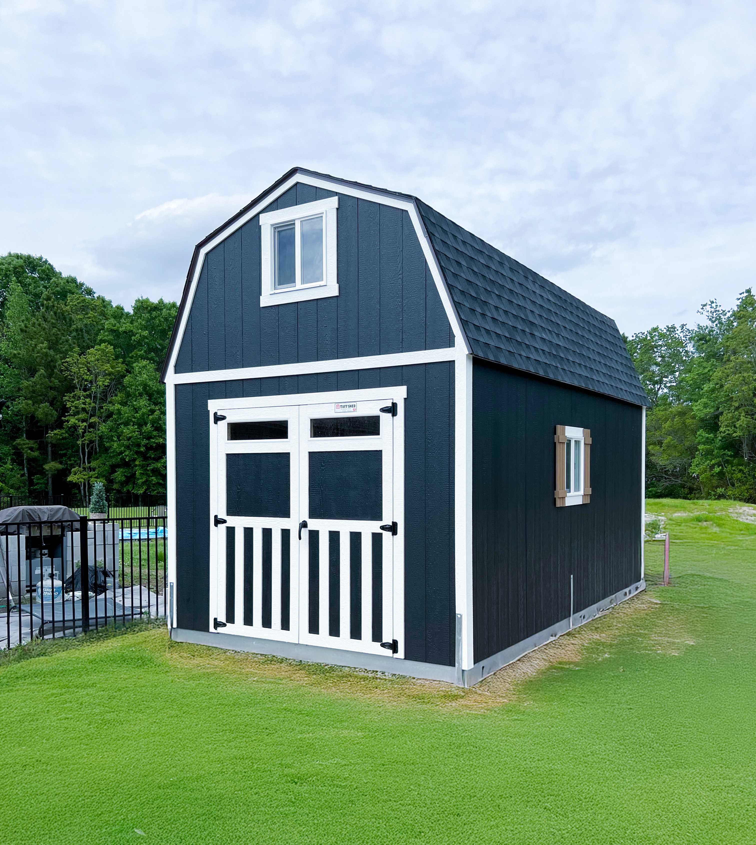 A dark blue barn-style shed with white trim and a gabled roof sits on a grassy area near trees and a fenced pool.