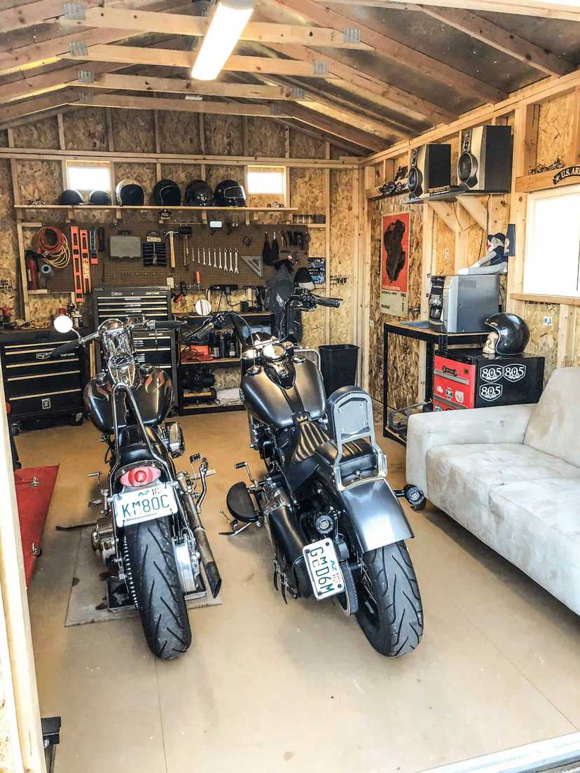 Garage interior with two motorcycles, a workbench with tools, a sofa, and storage shelves. Walls are unpainted wood, and a window provides light.