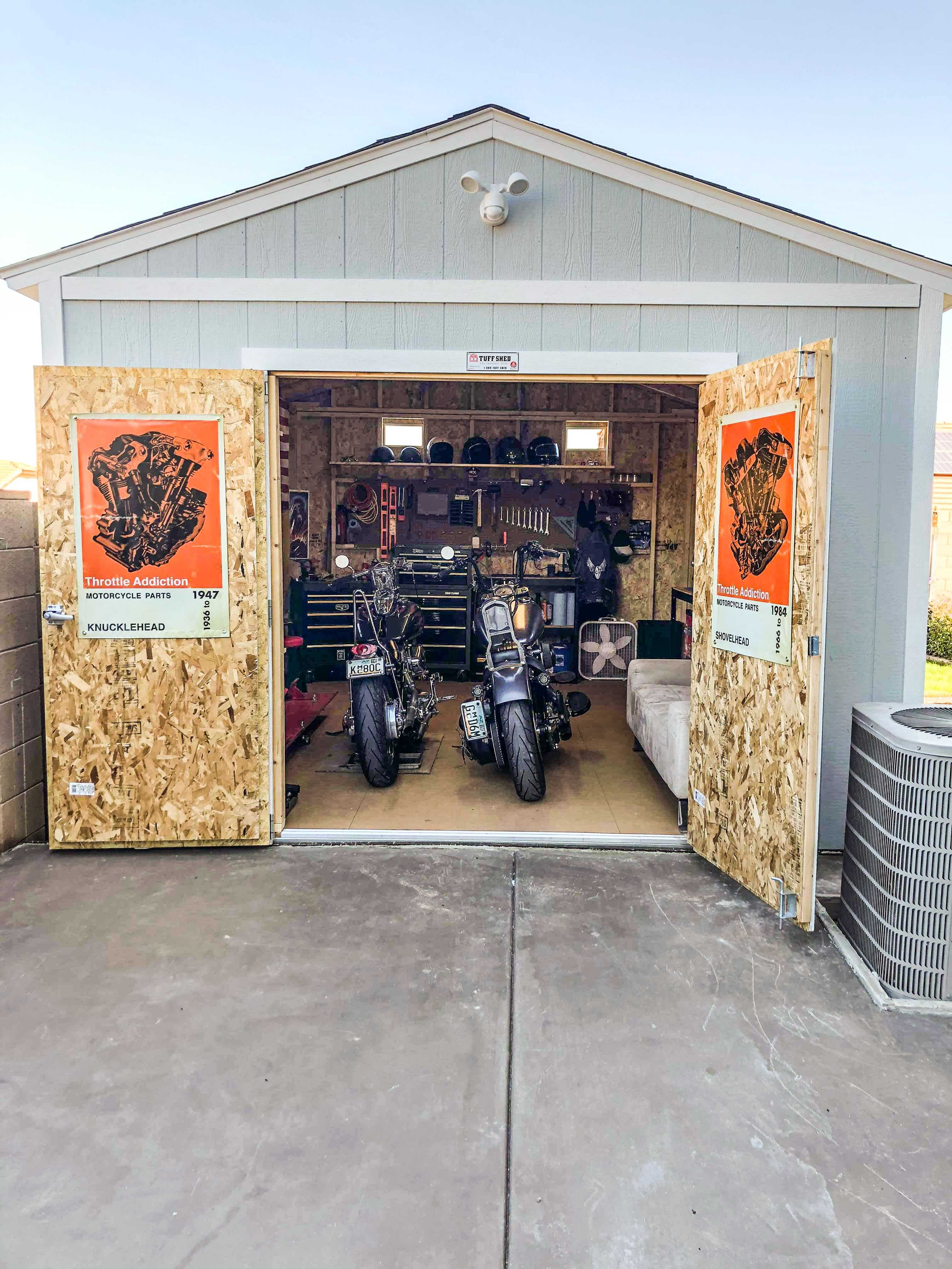 Open garage with two motorcycles inside, surrounded by tools, helmets, and posters on the doors.