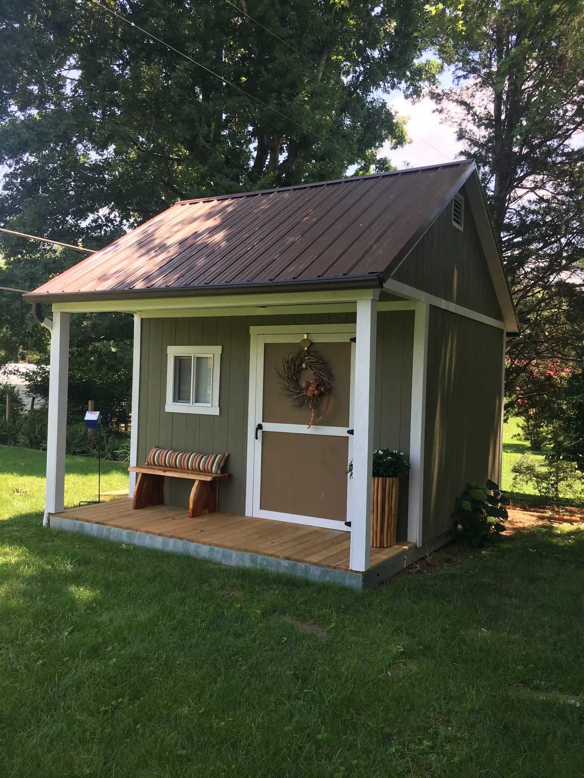 Small shed with a brown metal roof, gray walls, a porch with a bench, plants, and a door adorned with a wreath, surrounded by greenery.