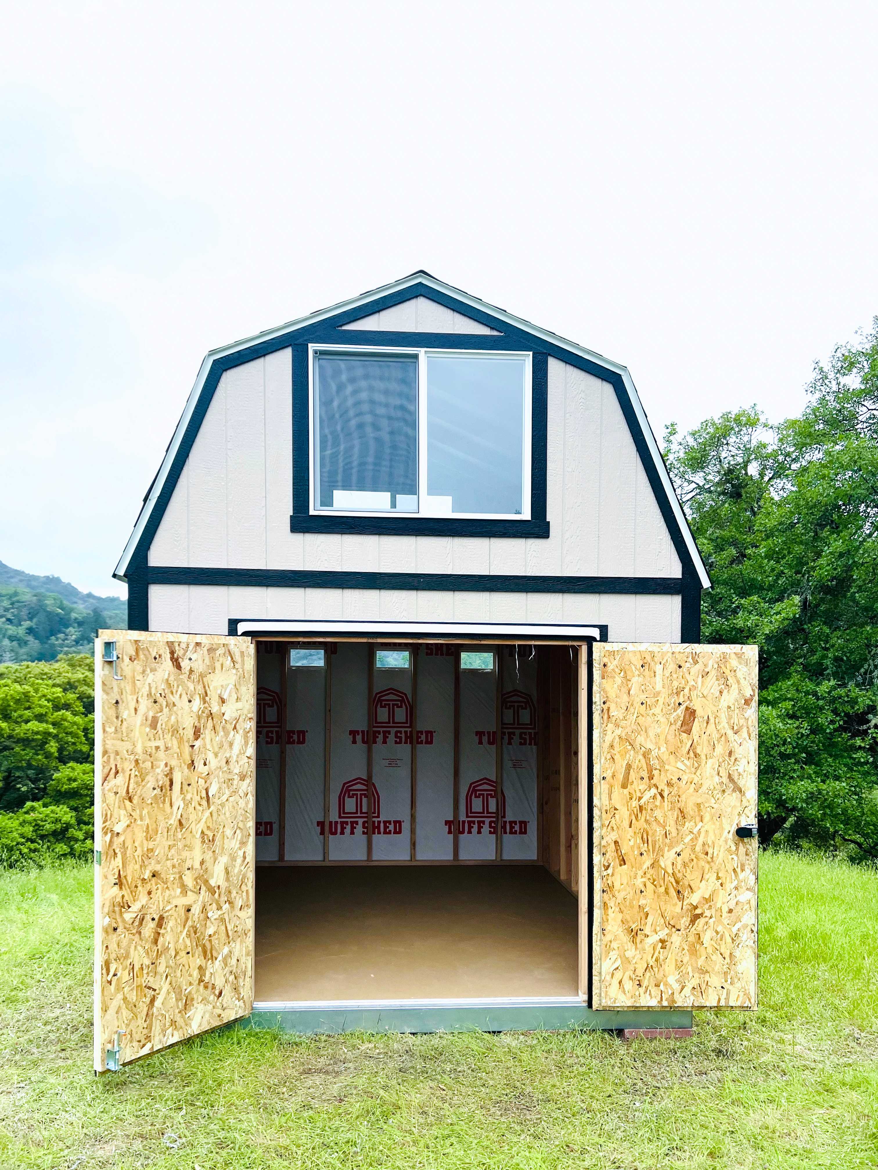 TB800 barn-style shed with open doors, revealing an unfinished interior. Set on a grassy field with trees and hills in the background.