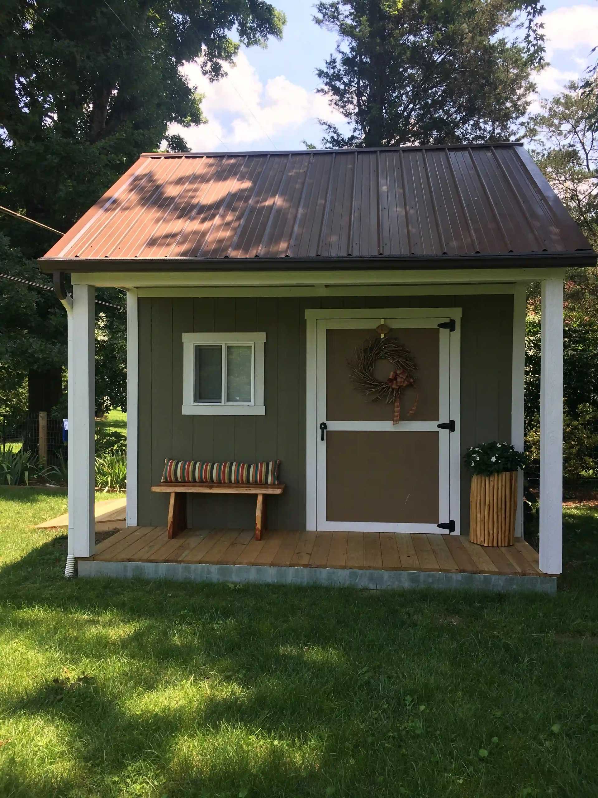 Small gray shed with a brown metal roof, front window, and door adorned with a wreath. Wooden bench and planter on the porch, surrounded by greenery.