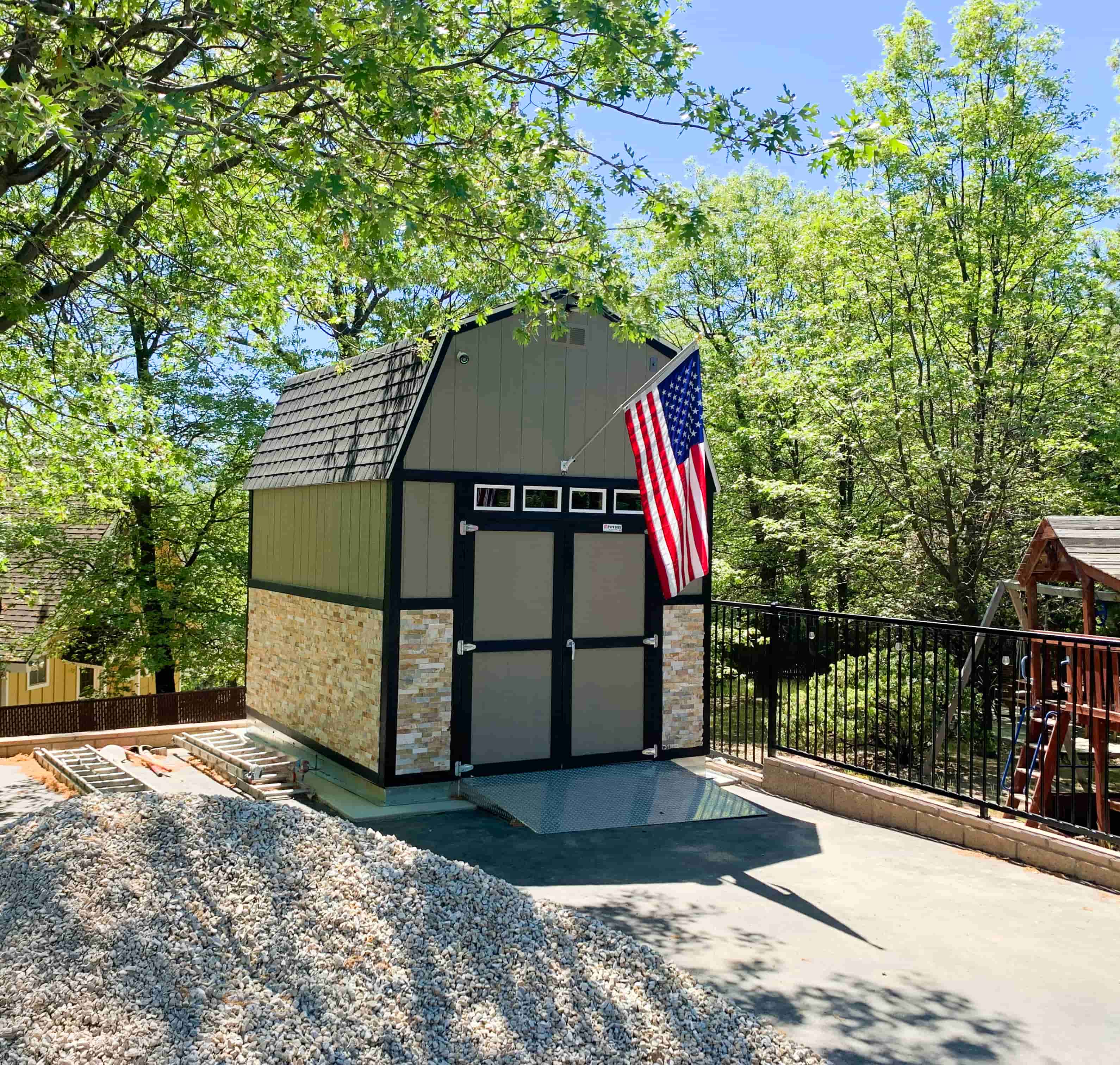 A barn-style shed with stone accents and an American flag. Surrounded by trees, with gravel and a playset nearby.