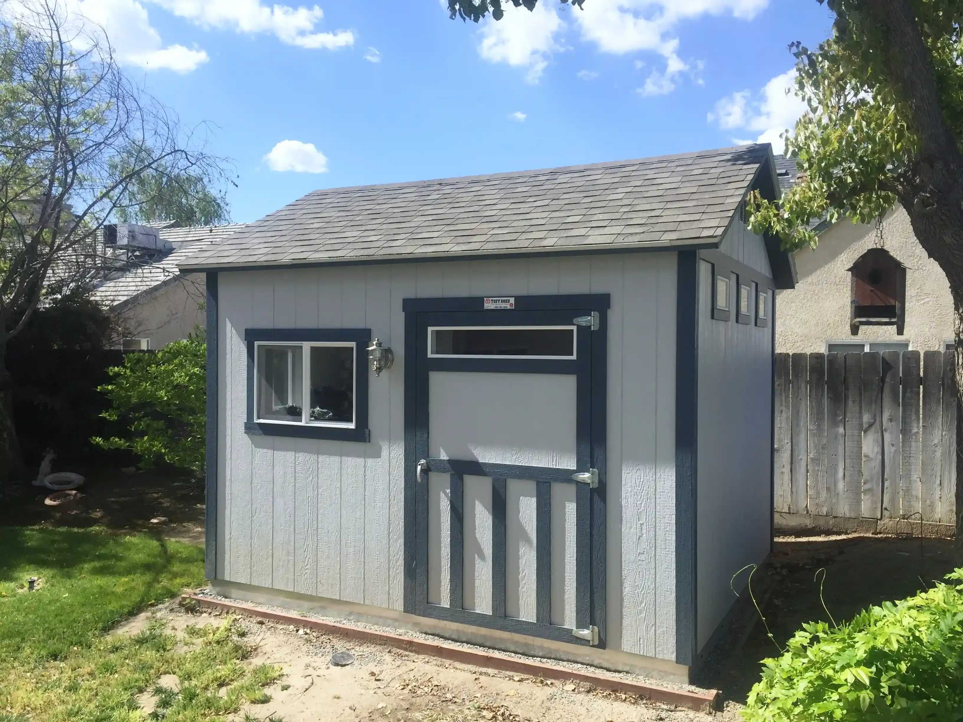 A small gray shed with a dark trim, two windows, and a door sits in a backyard with grass and trees under a blue sky