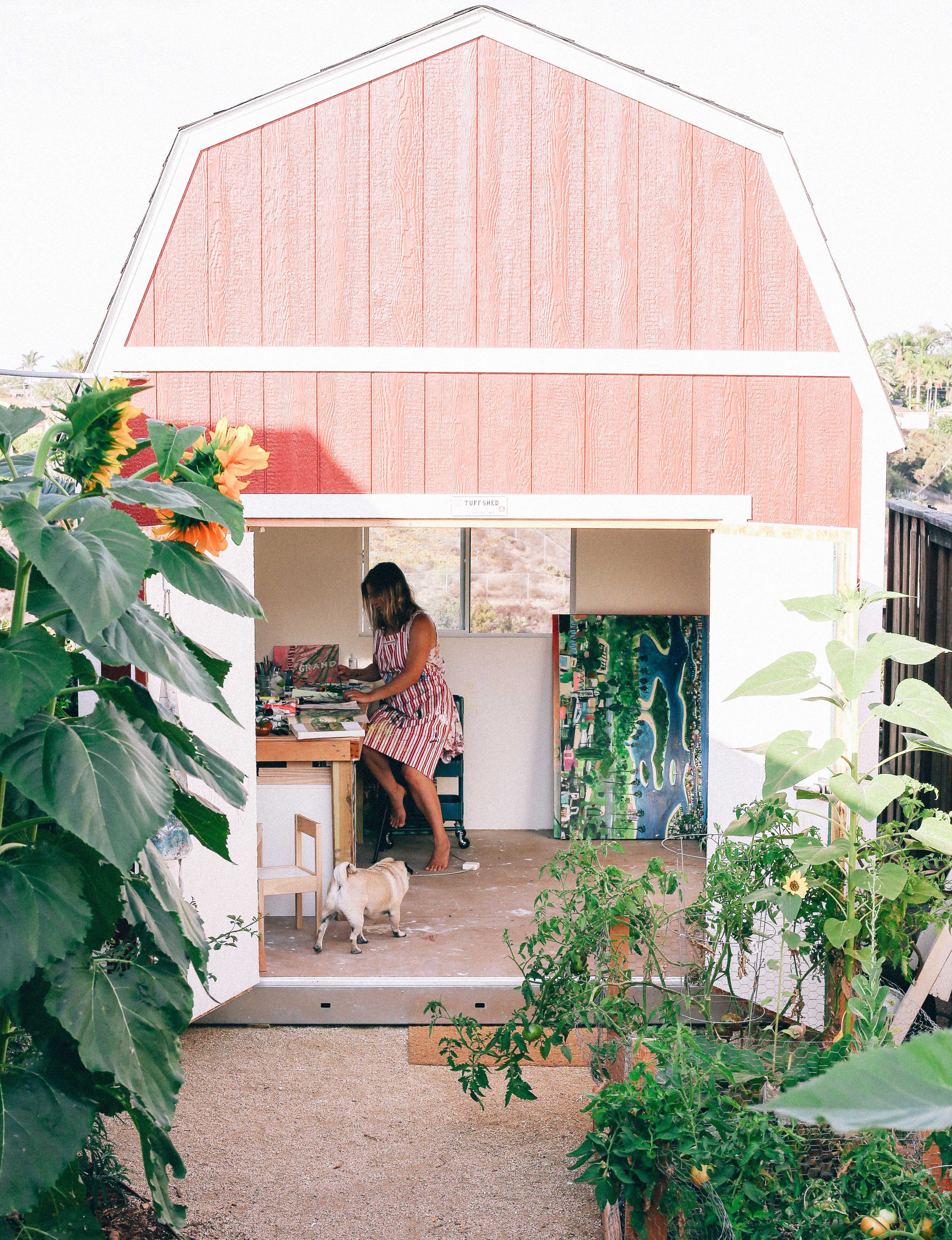 A woman in a striped dress works in a red TB700 shed studio, with a dog nearby and large sunflowers and plants surrounding the area.