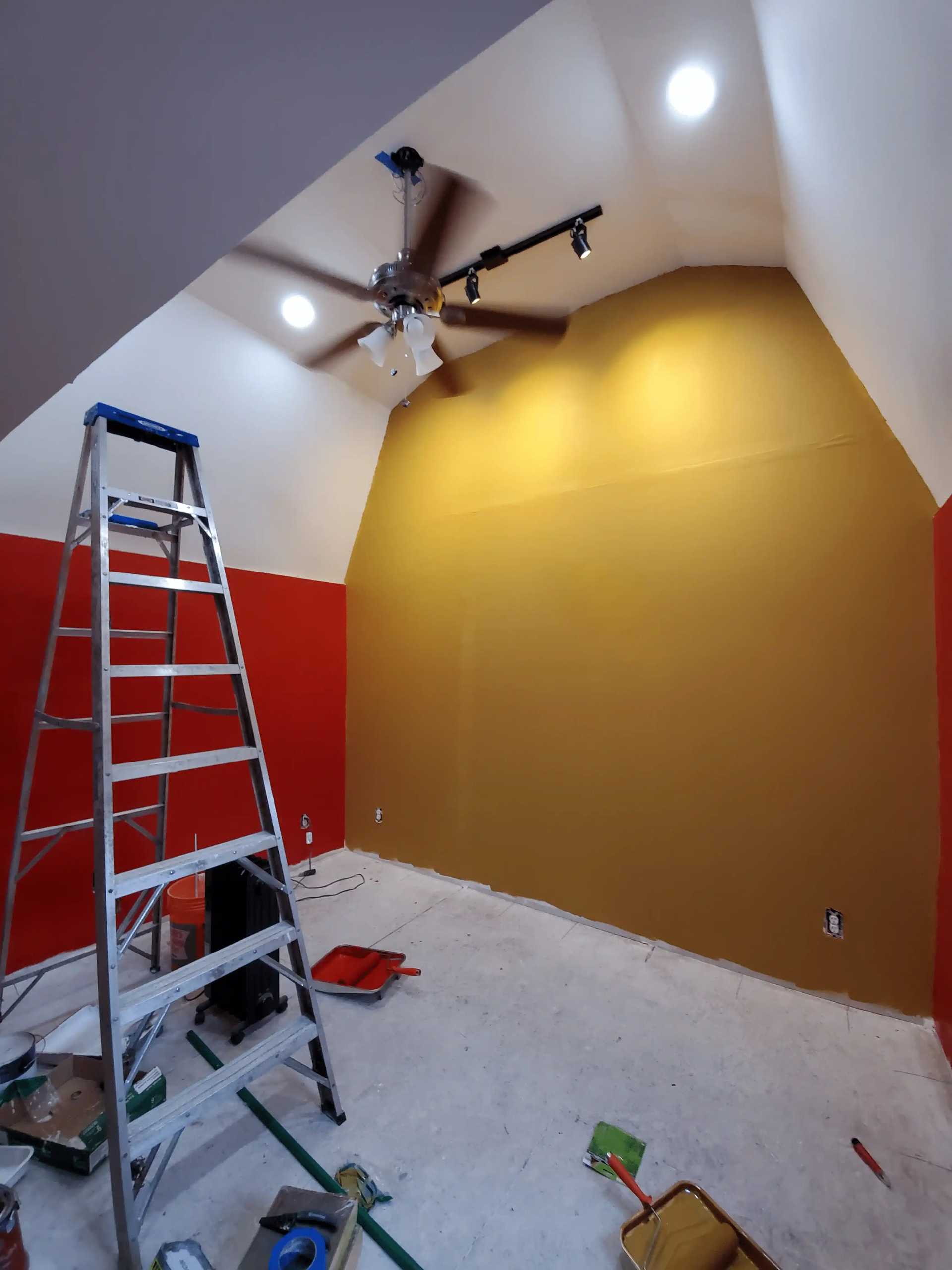 Room with a high ceiling, red and yellow painted walls, ceiling fan with lights, ladder, and painting supplies on the floor.