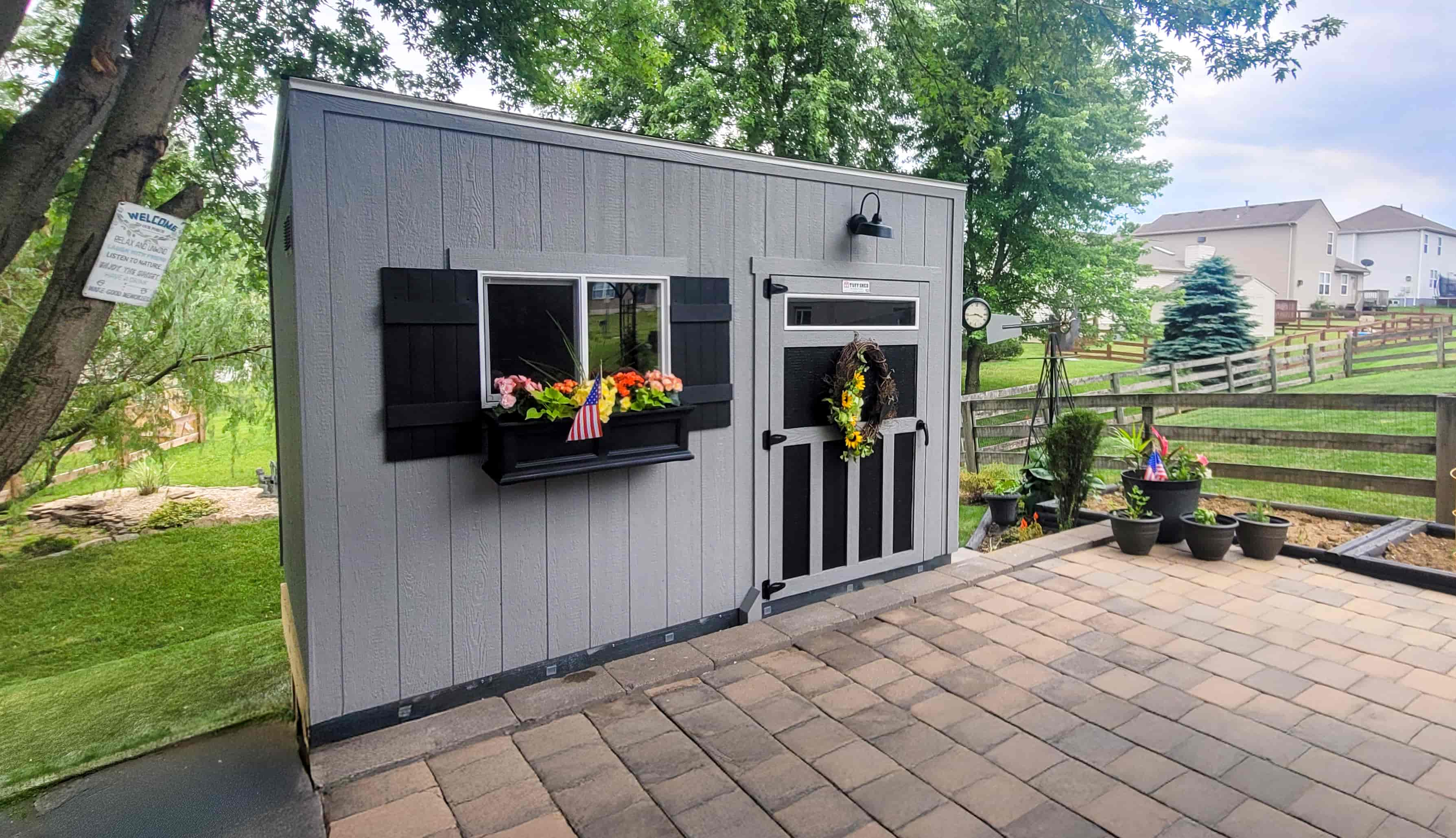 A gray garden shed with black shutters, adorned with flowers and a wreath, stands on a paved patio in a suburban backyard.