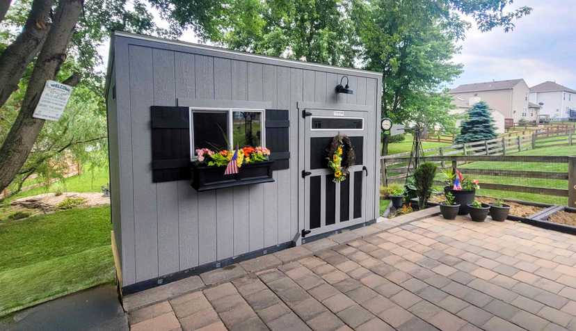 A gray garden shed with black shutters, adorned with flowers and a wreath, stands on a paved patio in a suburban backyard.