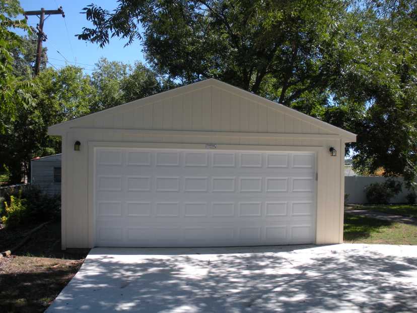 Beige two-car garage with white doors, surrounded by trees and greenery, under a clear blue sky.