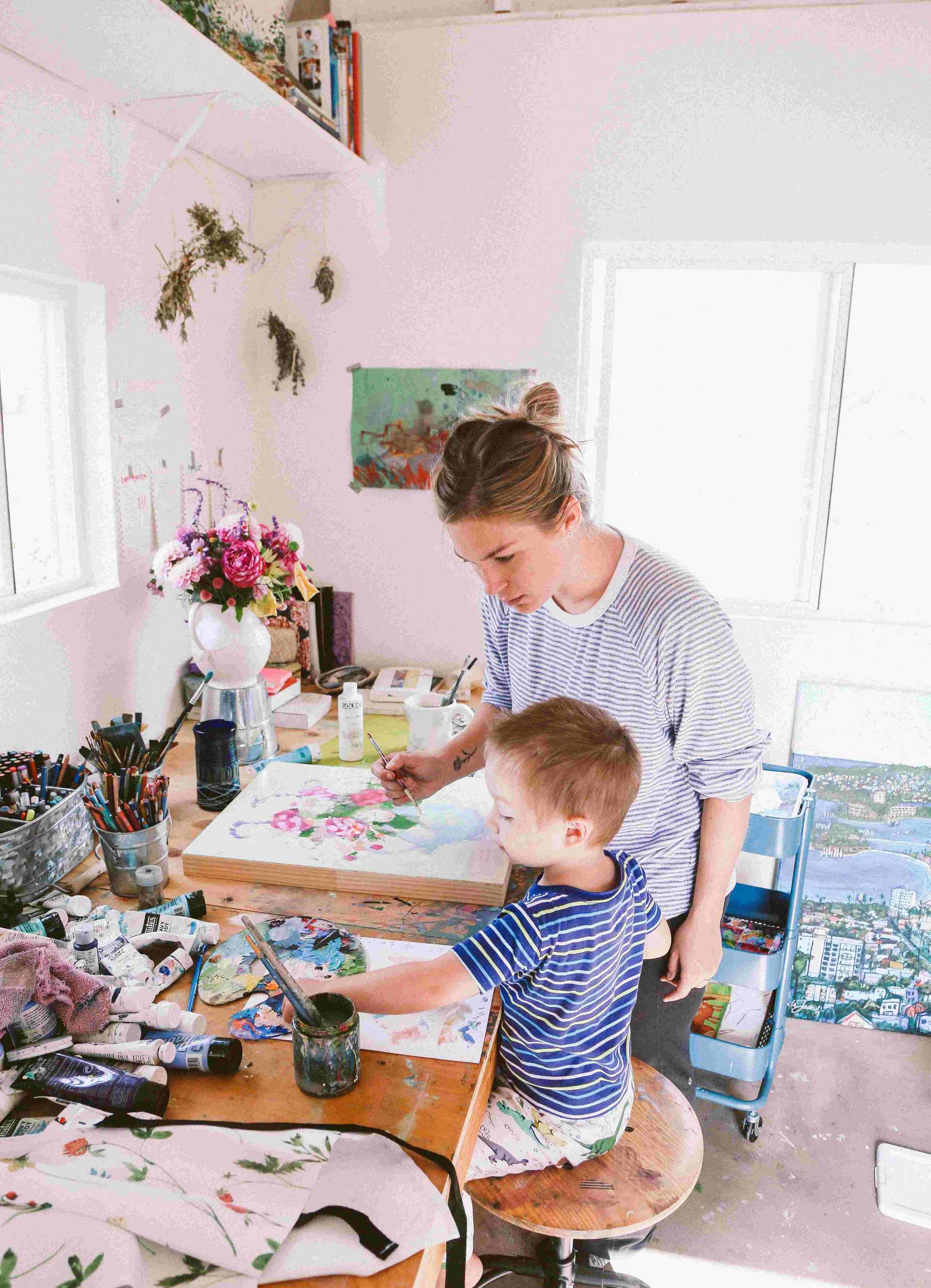 Michaela Jean, the artist at work, paining a bouquet of flowers with her son painting next to her. Tje are at the art studio, a red tuff shed barn