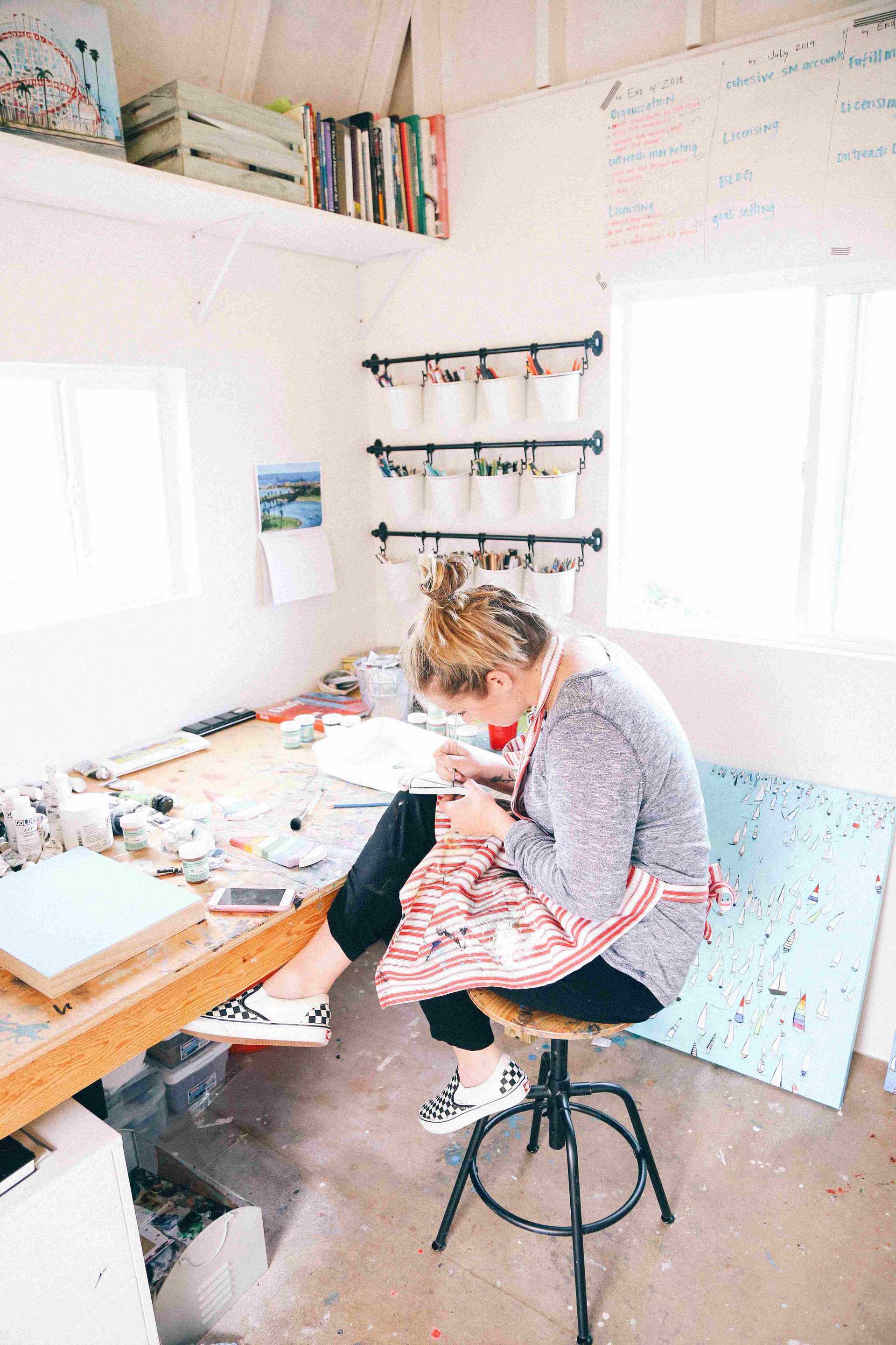 an artist at work, seating on the round stool and sketching. With canvases by the wall, books and pencils
