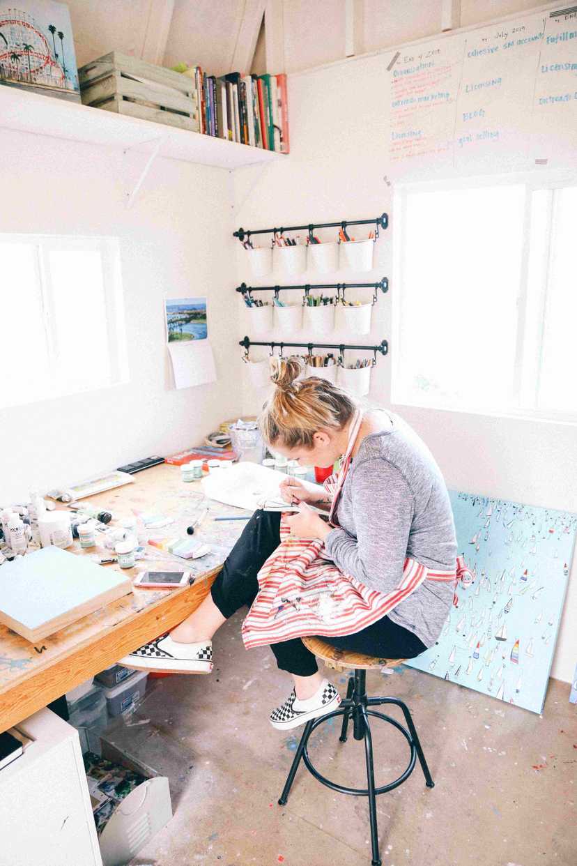 an artist at work, seating on the round stool and sketching. With canvases by the wall, books and pencils