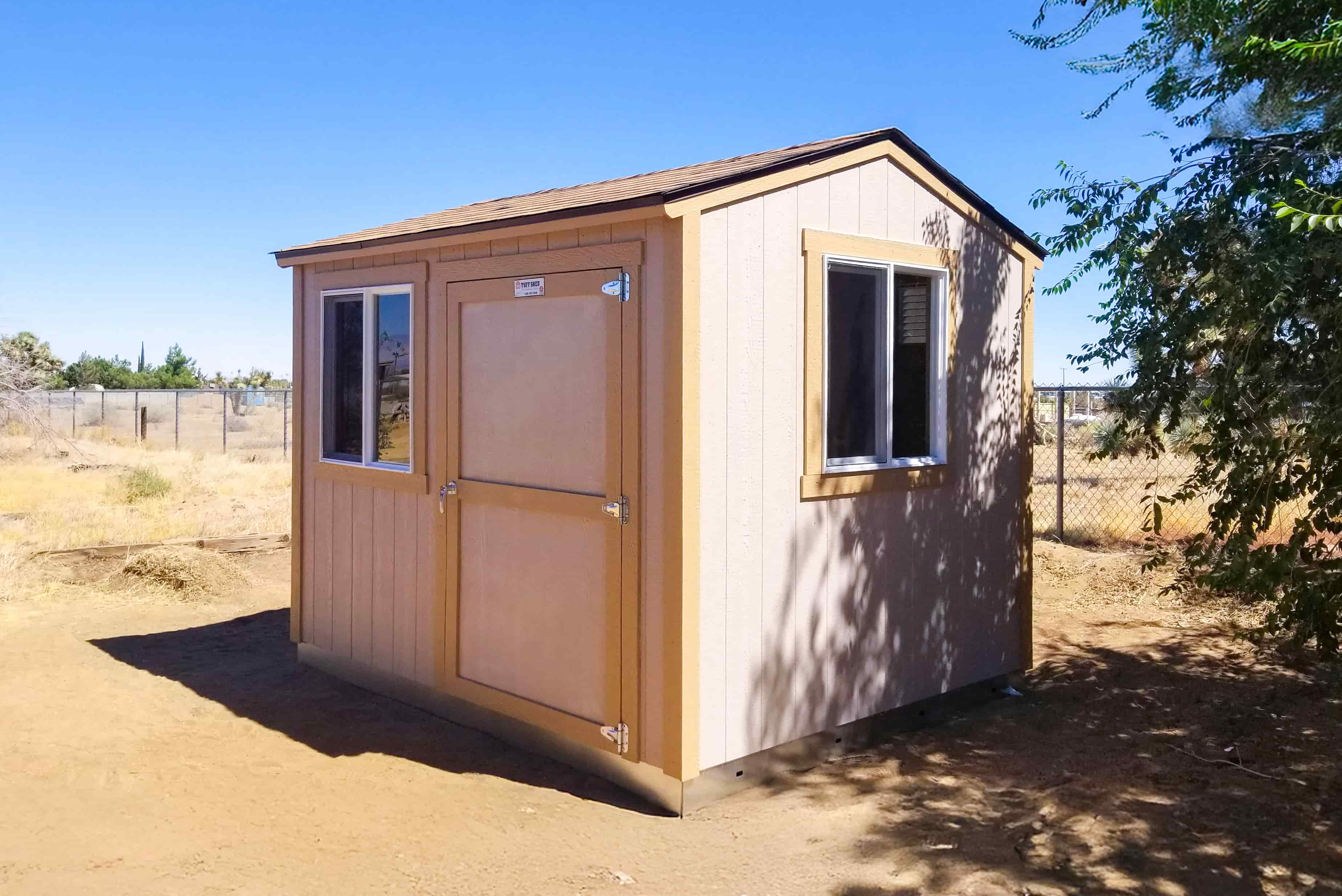 A TR700 beige and brown shed with a window and door stands on dry, sandy ground, surrounded by a fence and sparse trees under a clear blue sky.