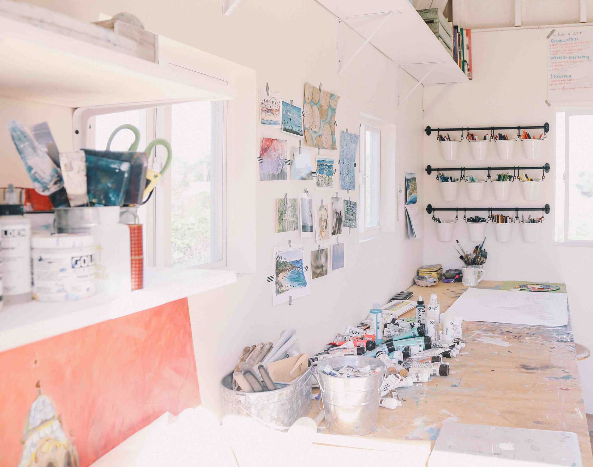 an interior shot of the artist studio with workbench table set along the wall by the windows, with art inspiration on the wall, buckets of color pencils and paint tubes