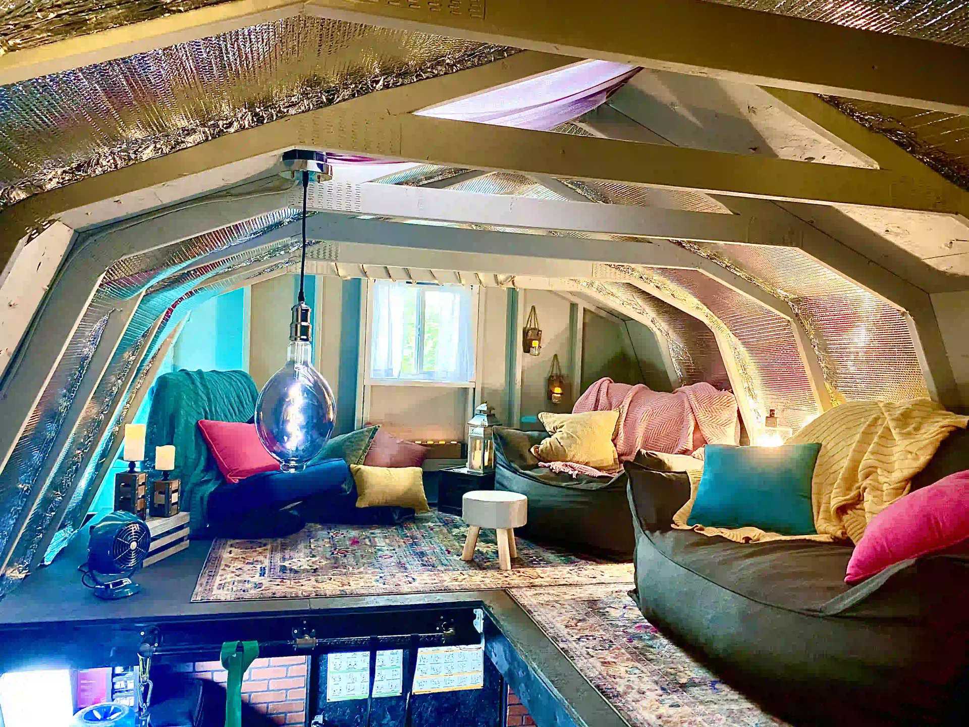 Cozy attic space with bean bags, colorful pillows, a patterned rug, and ambient lighting. A window lets in natural light.