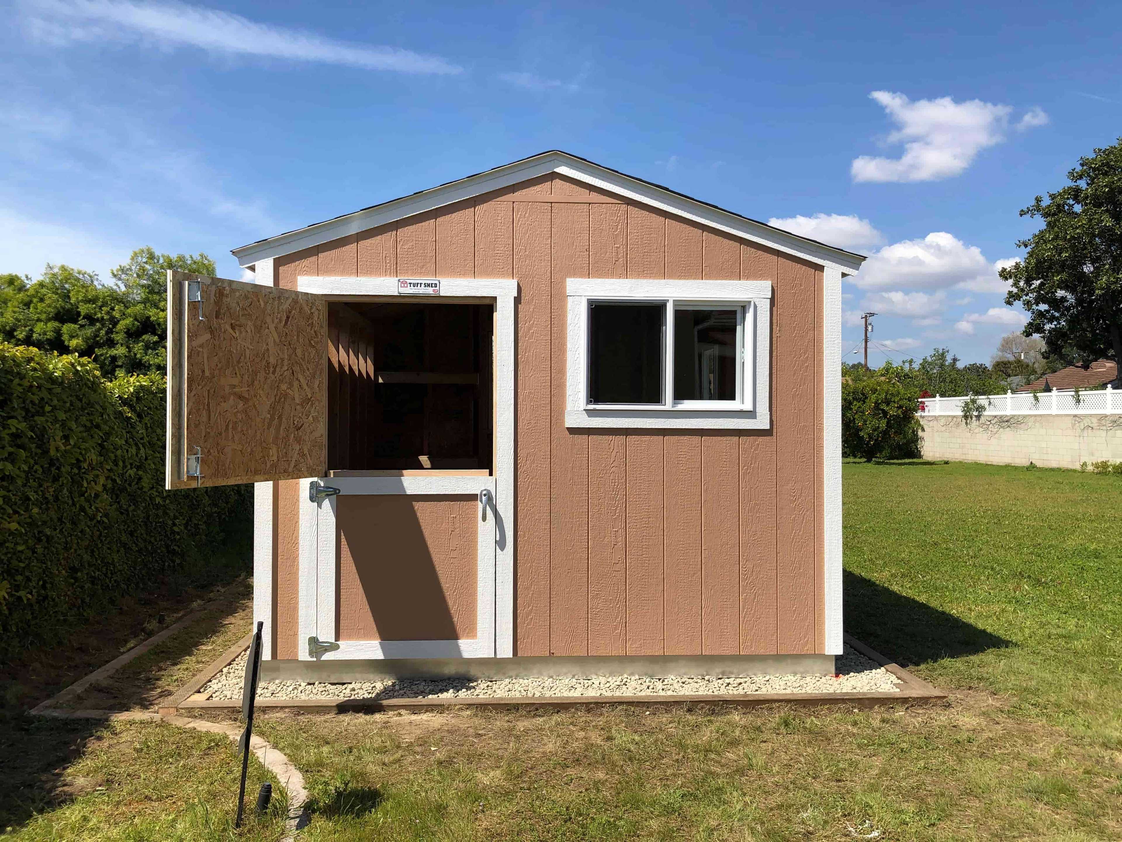 A small, brown wooden shed with white trim and multiple windows sits on a grassy lawn, surrounded by a hedge and trees under a blue sky.