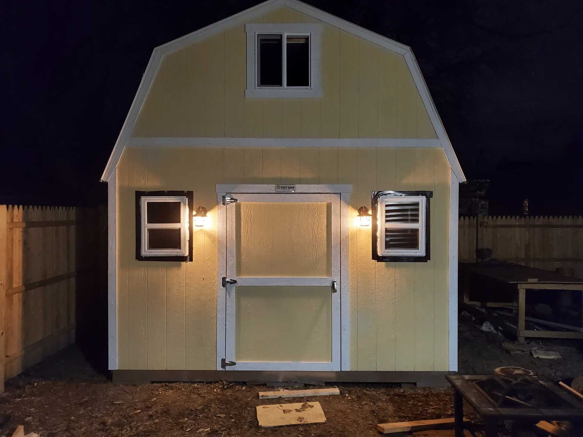 Yellow barn-style shed illuminated at night, featuring a central door with windows and exterior lights on each side, surrounded by a wooden fence.