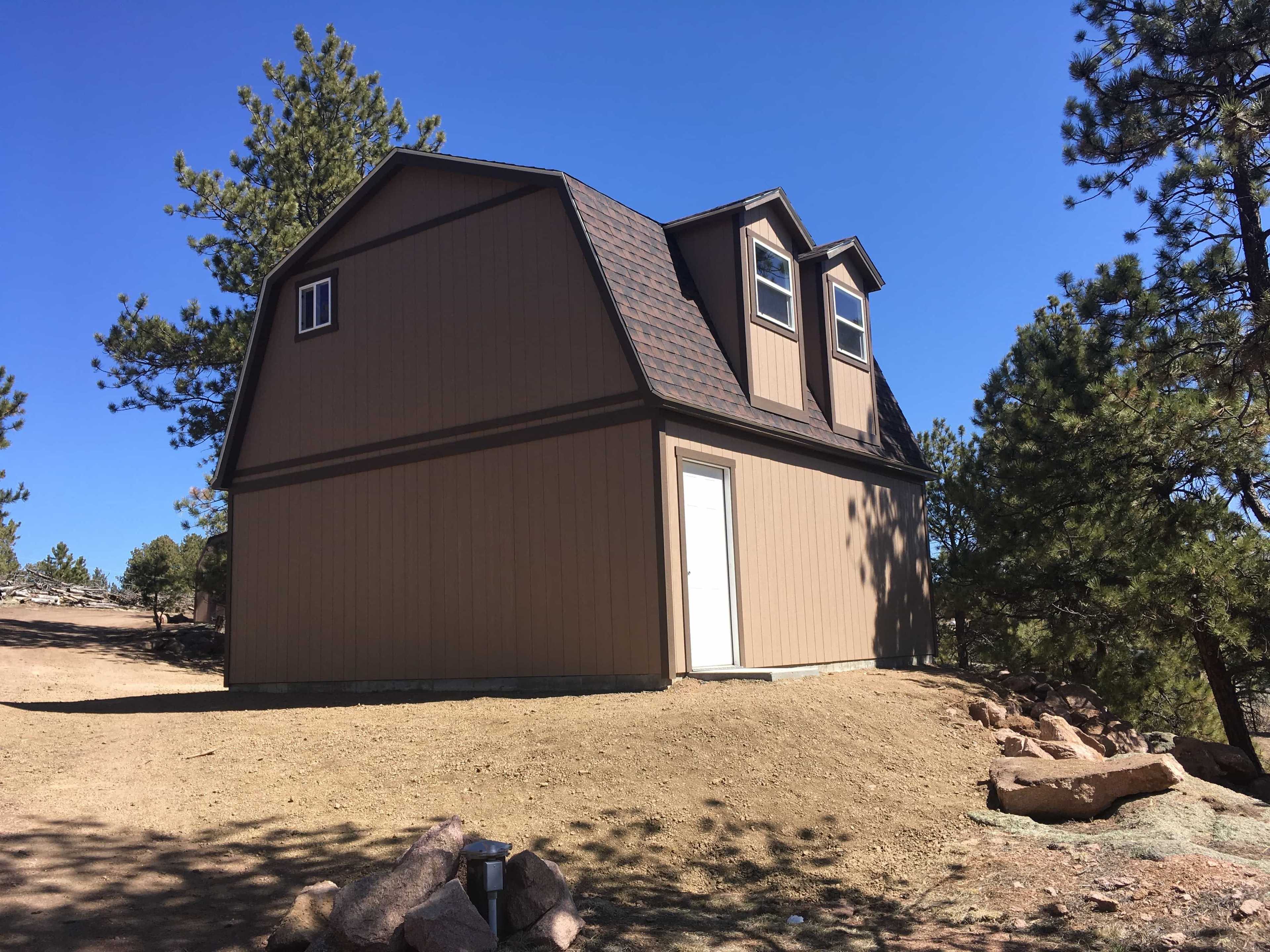 A small, tan, barn-style house with a white door sits on a dirt hill surrounded by pine trees under a clear blue sky.
