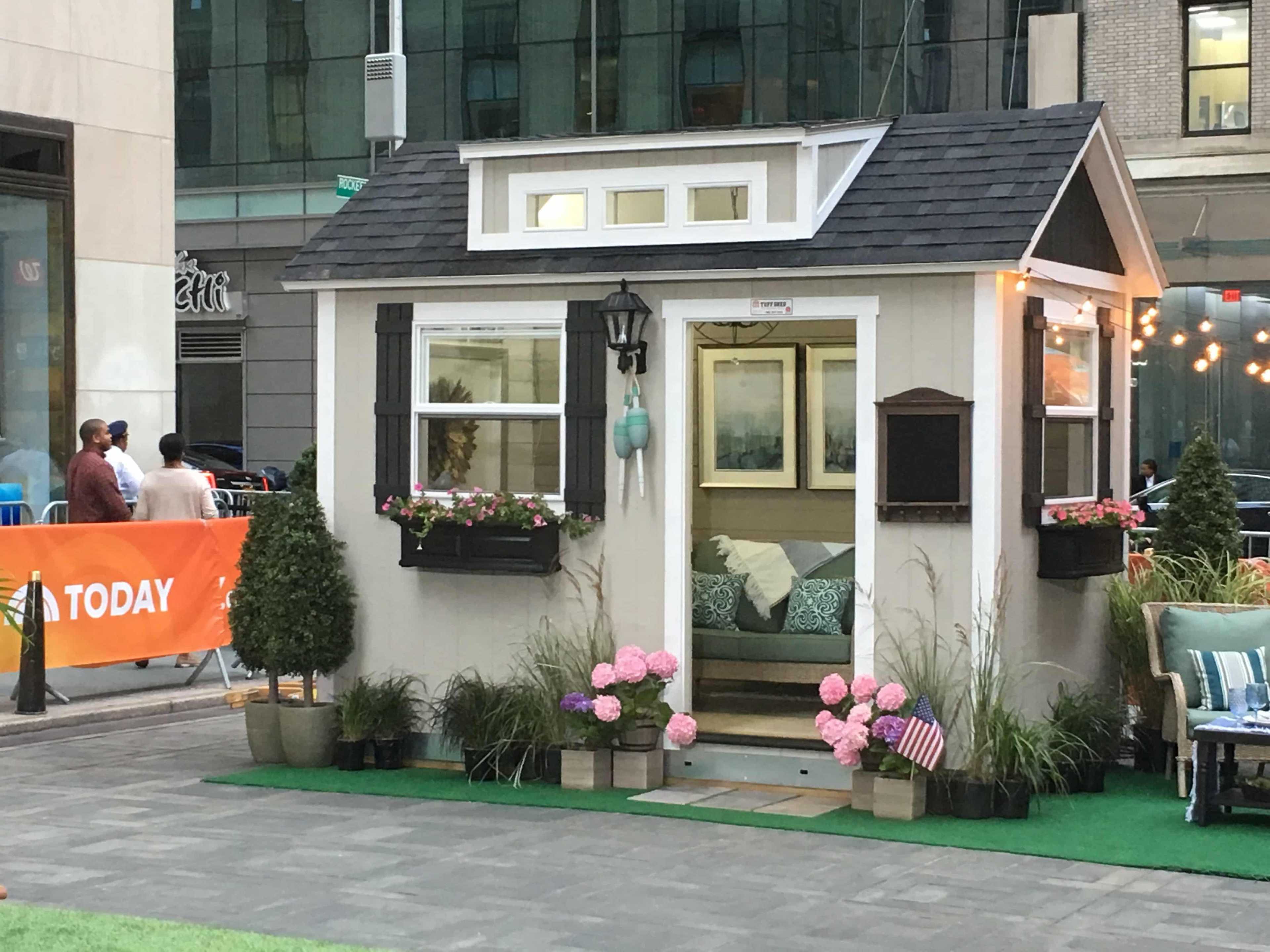 A small, charming house with flower boxes and a tiny porch, set in an urban area with modern buildings in the background.