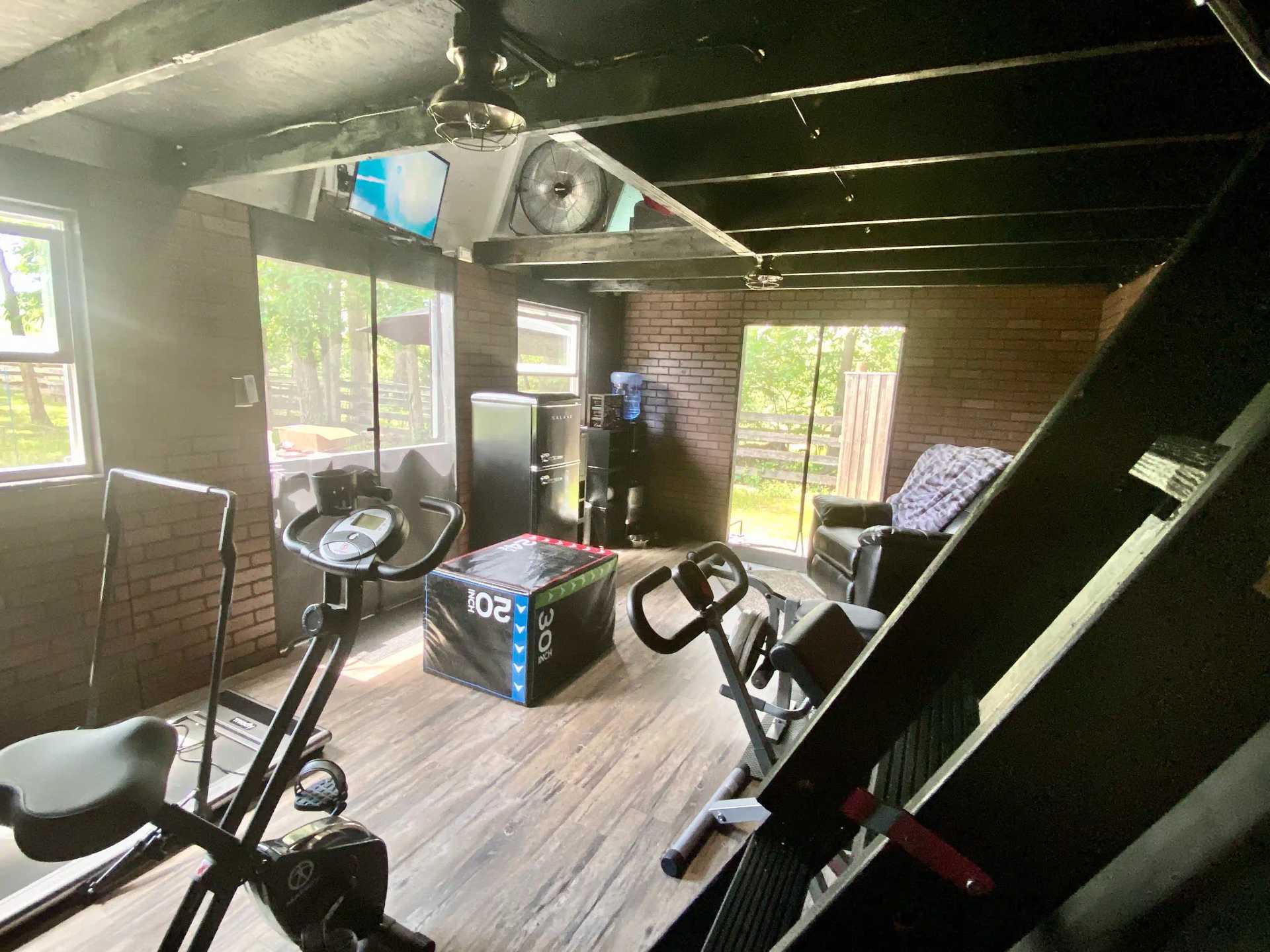 Home gym with exercise bikes, a treadmill, a rowing machine, and a weight box. Brick walls and windows showing greenery outside.