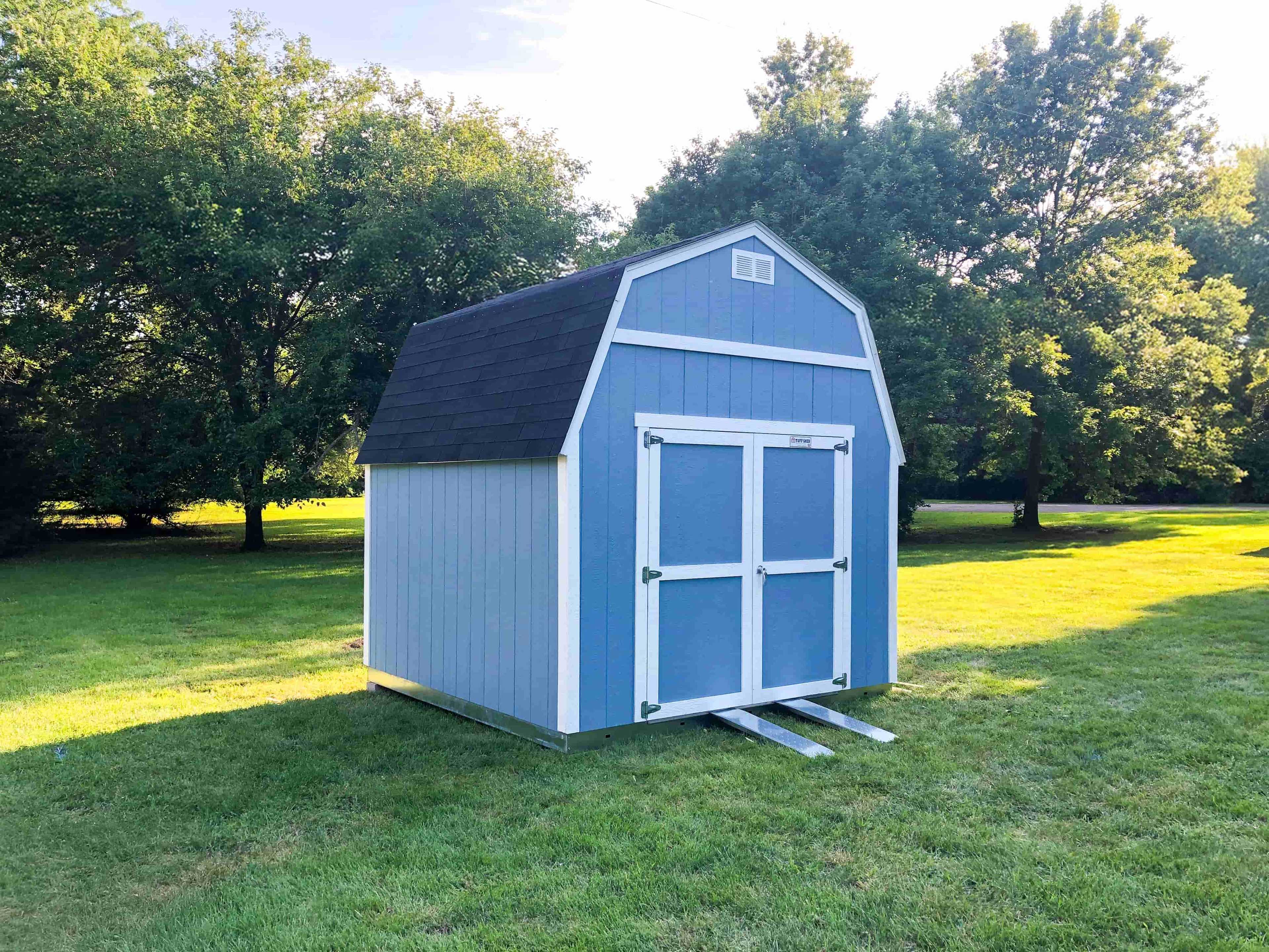 A TB600 blue shed with white trim and a gabled roof sits on a grassy lawn, surrounded by trees.