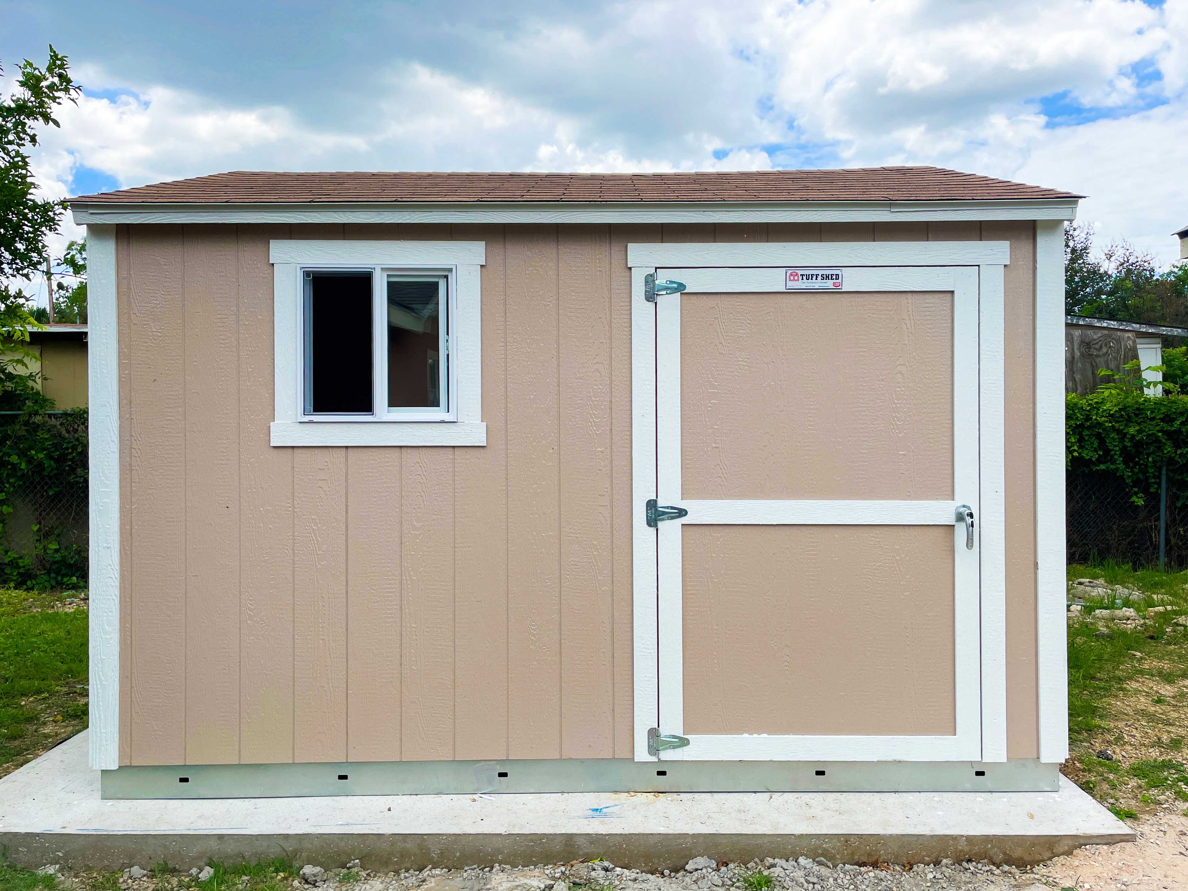 A TR700 beige shed with a sloped brown roof, a window on the left, and a white door on the right, set on a grassy area under a cloudy sky.
