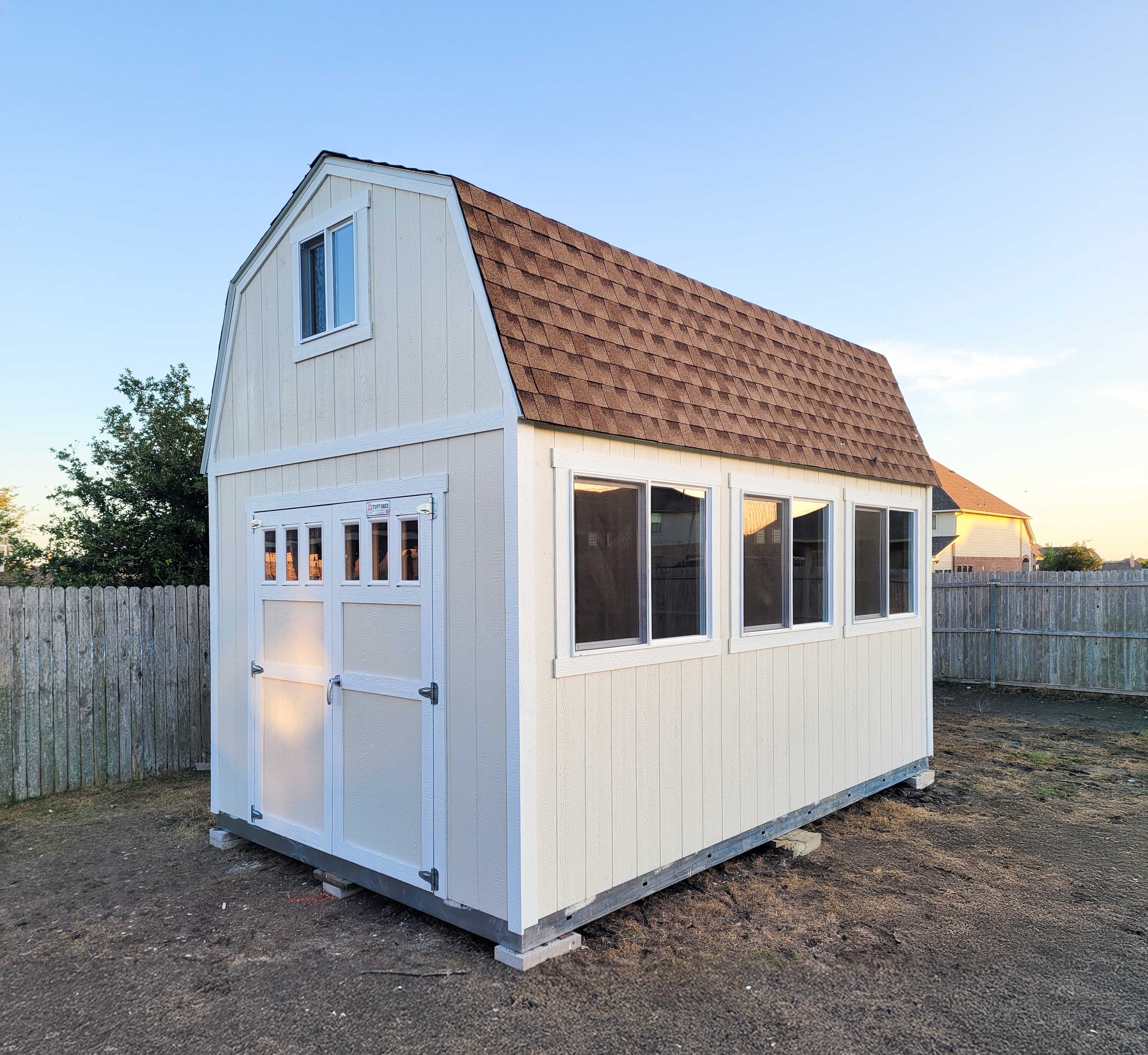 A beige shed with a brown shingled roof and multiple windows stands on a dirt patch, surrounded by a wooden fence.