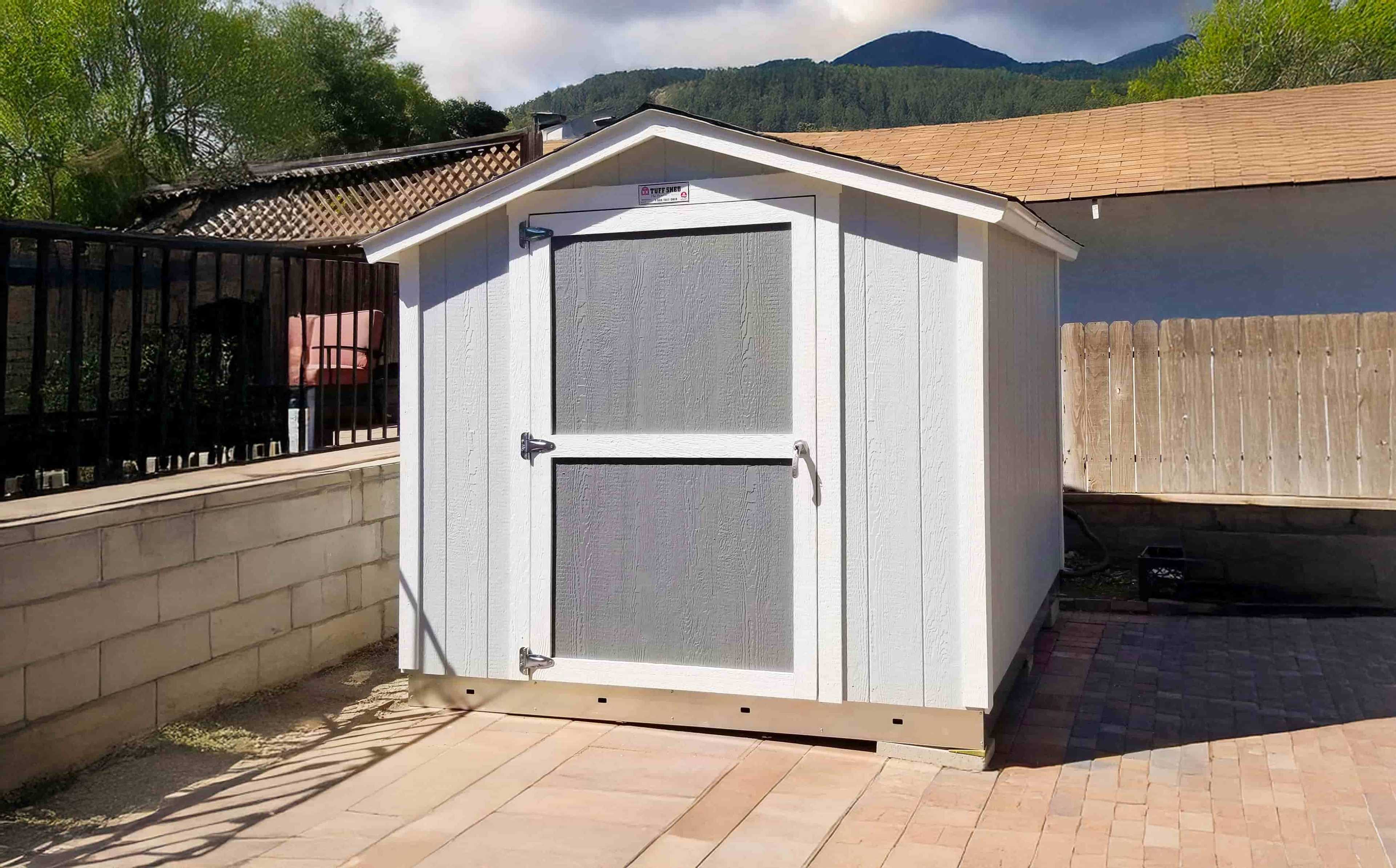 A white SR600 wooden shed with a door stands on a paved area, surrounded by a wooden fence
