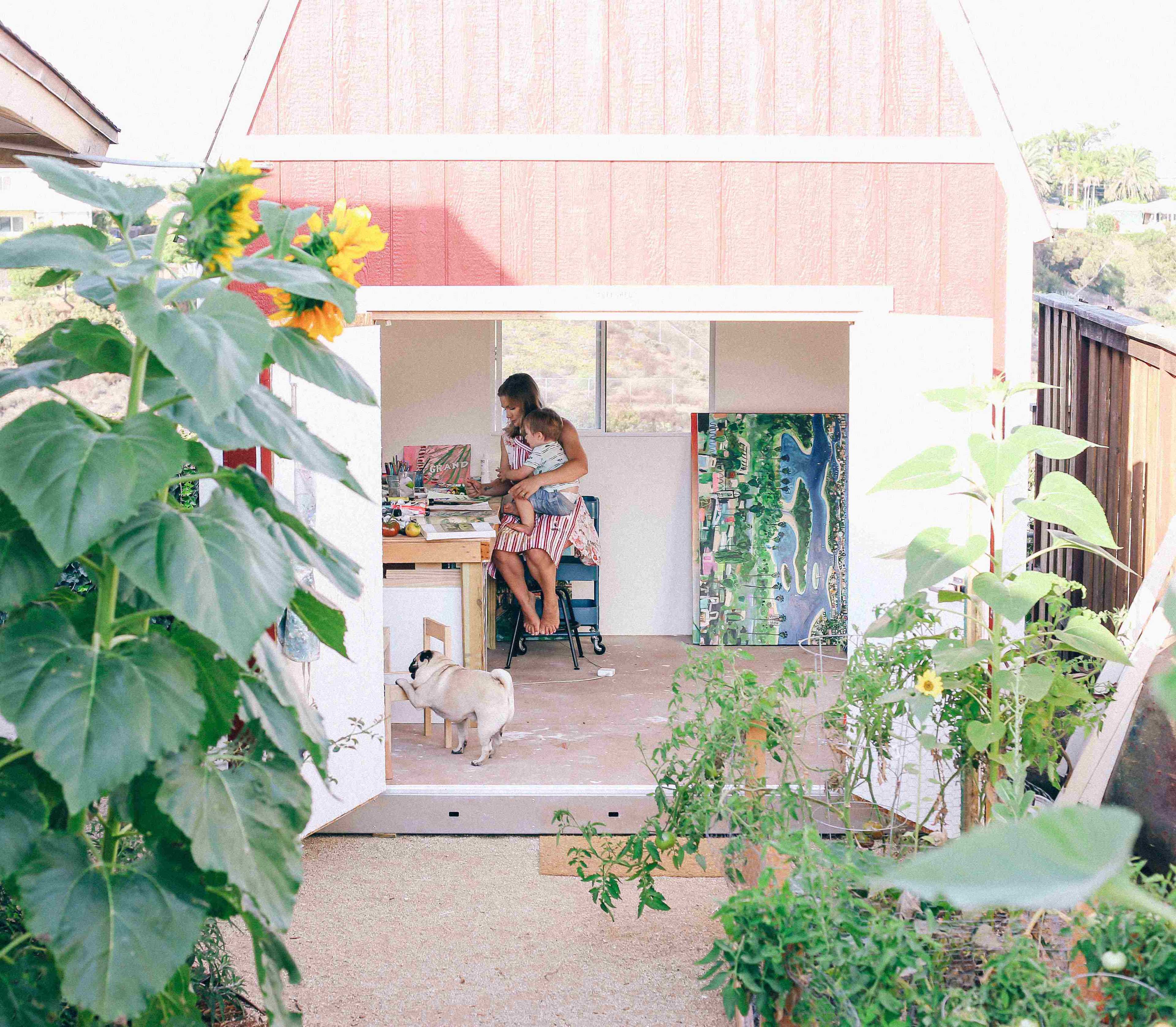 Michaela Jean, the artist holding a child on her lap, at her art studio–a red tuff shed barn set in the blooming garden with wide open double doors.