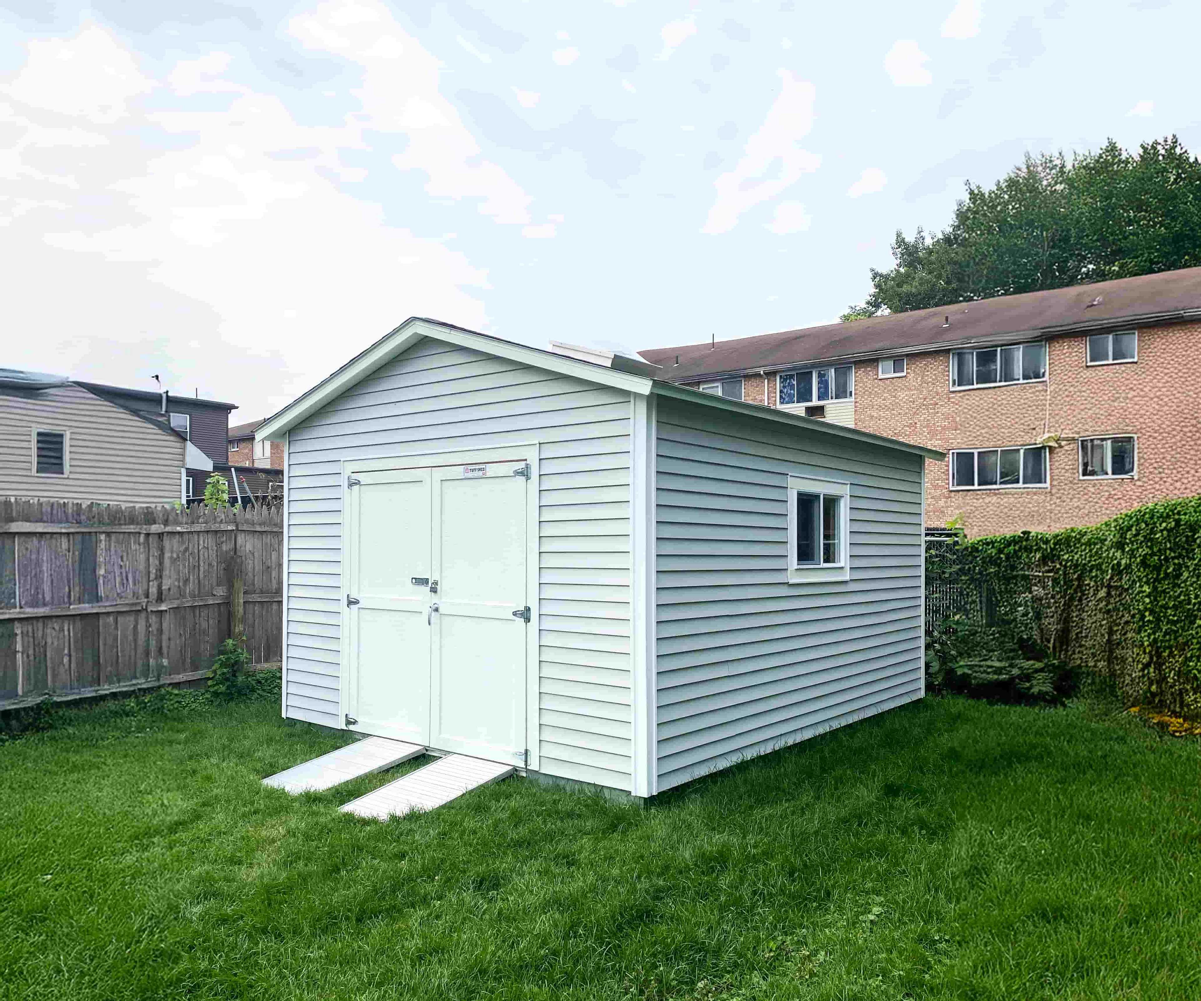 A TR800 light gray shed with white trim and a ramp stands on a grassy lawn, surrounded by a wooden fence and neighboring houses.