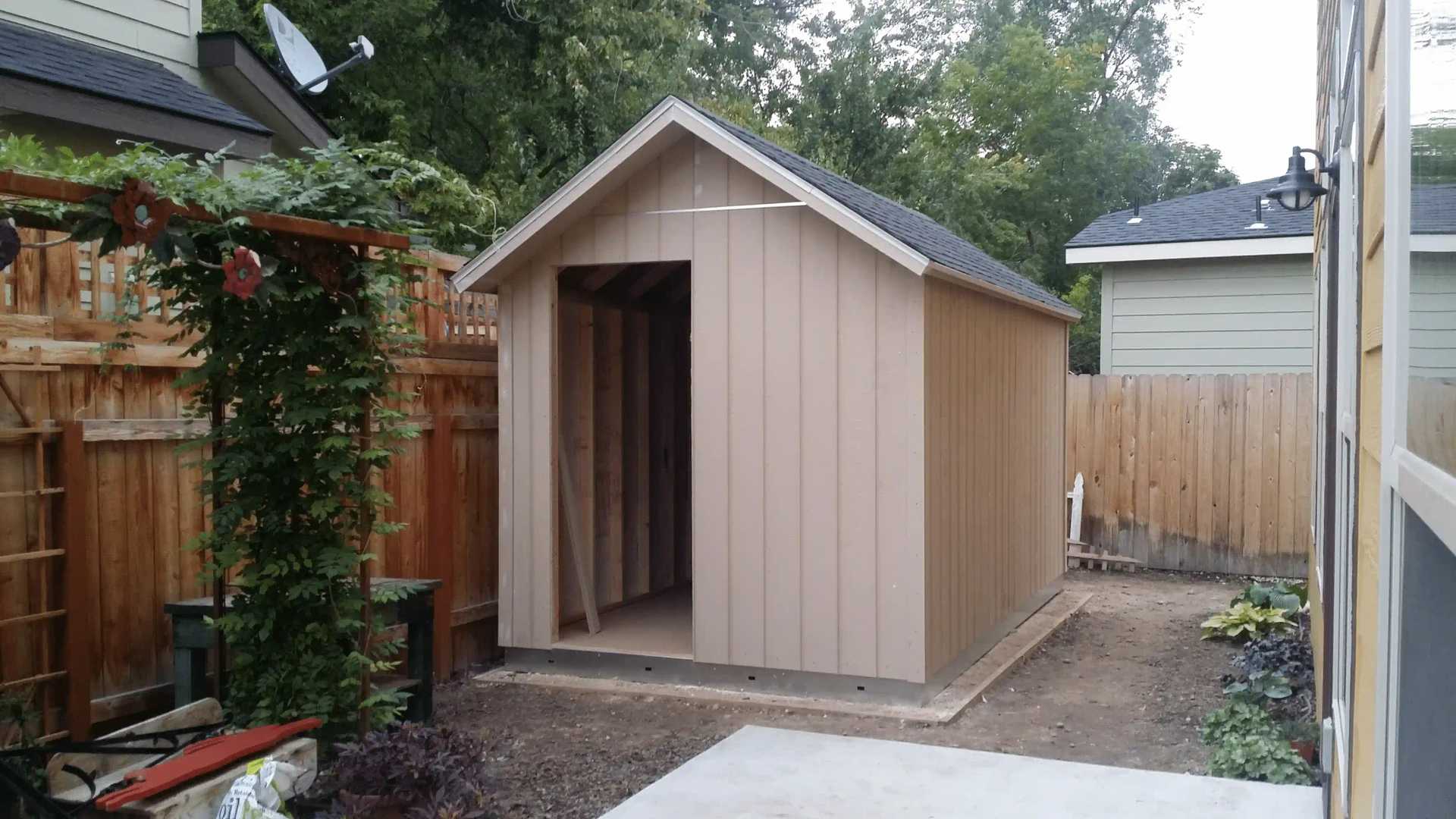 A small, unfinished wooden shed with an open entrance, situated in a fenced backyard with surrounding trees and plants.